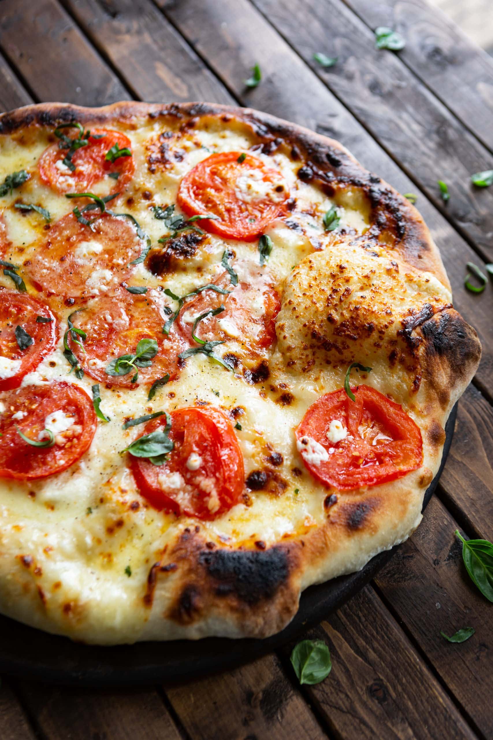 a photo of a whole four cheese margherita pizza with slices of tomato on top, melted golden cheese, chopped basil and chunks of feta cheese.