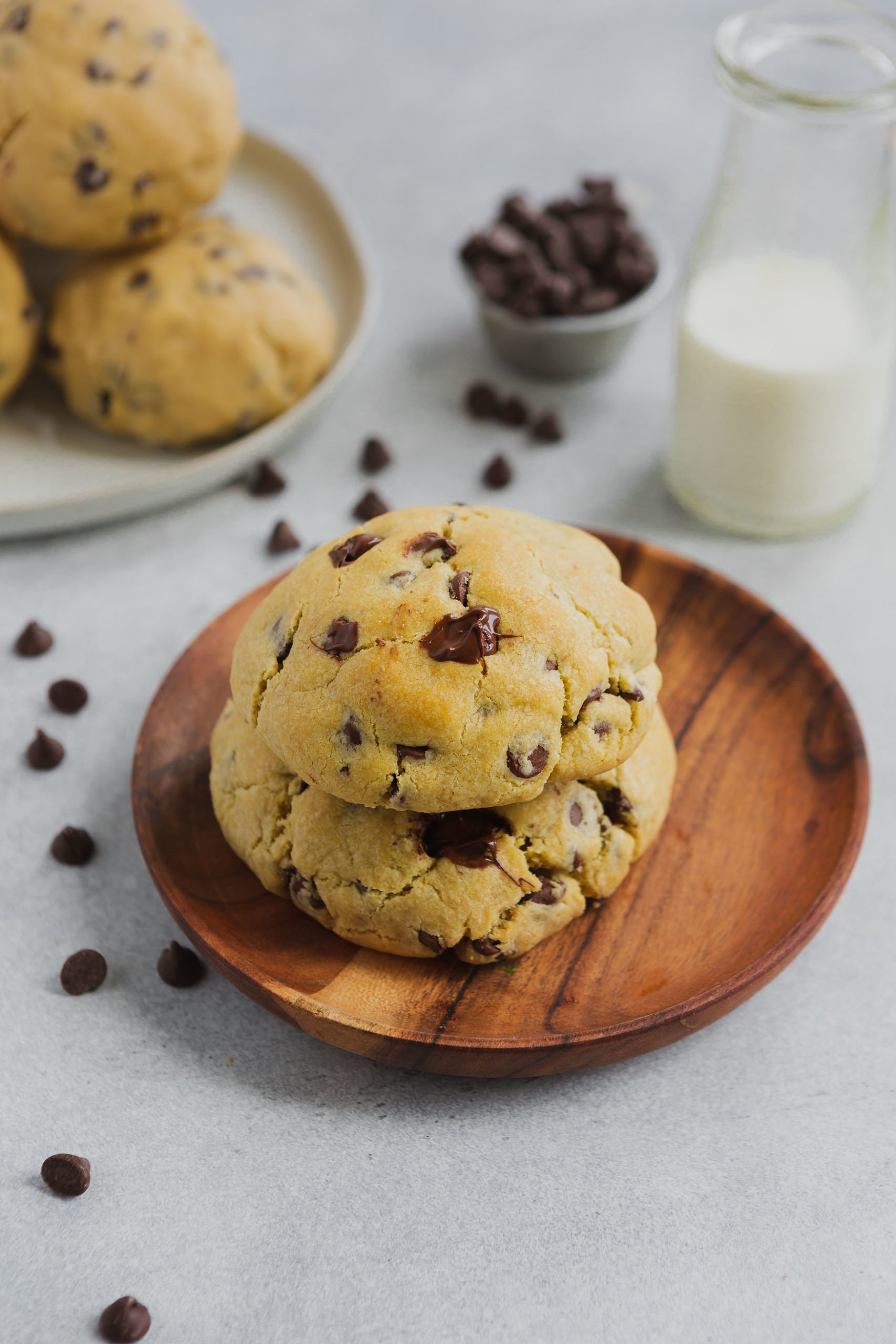 a photo of two large and thick chocolate chip cookies stacked on each other on a small wooden plate with a plate of more cookies in the background and a small cup or chocolate chips and a carafe of milk.
