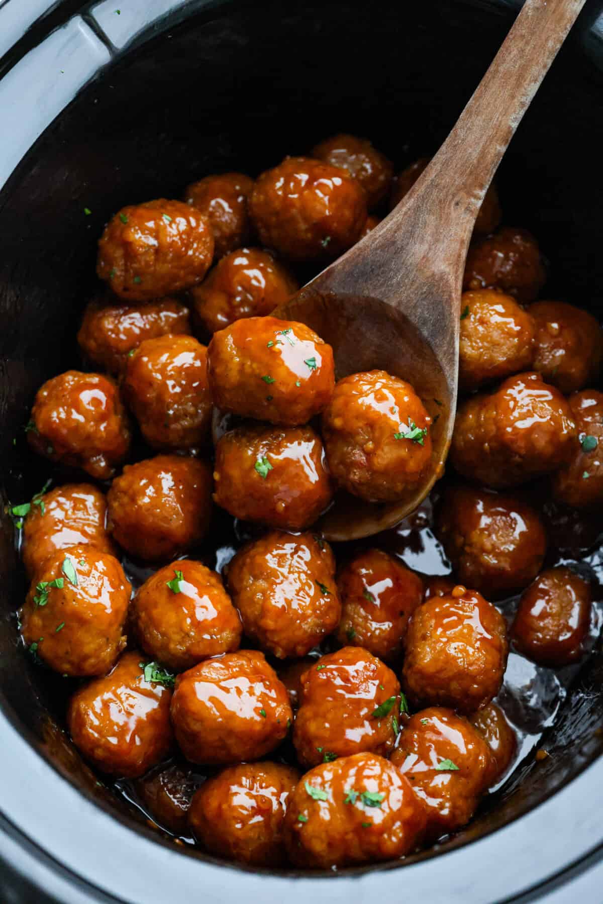 Someone taking a large wooden spoon and scooping a few slow cooker bourbon meatballs out of the crockpot.