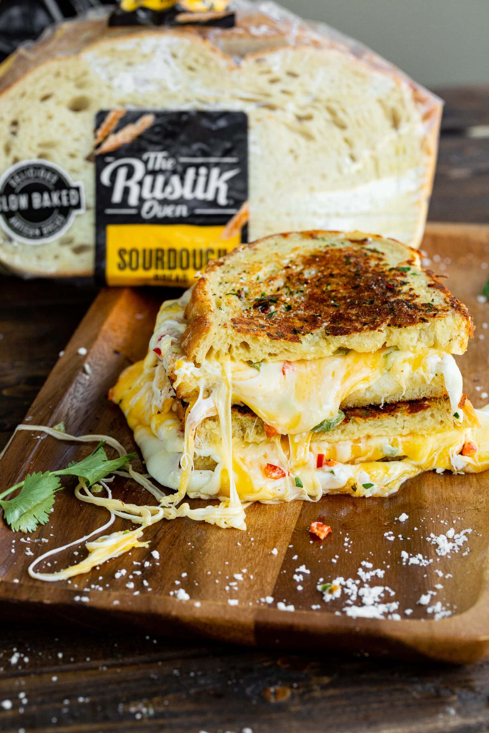 a photo of a grilled cheese sandwich that is crispy and golden on the outside with melted cheese dripping out of the inside of the sandwich with a bag of The Rustik Oven sourdough bread in the background.