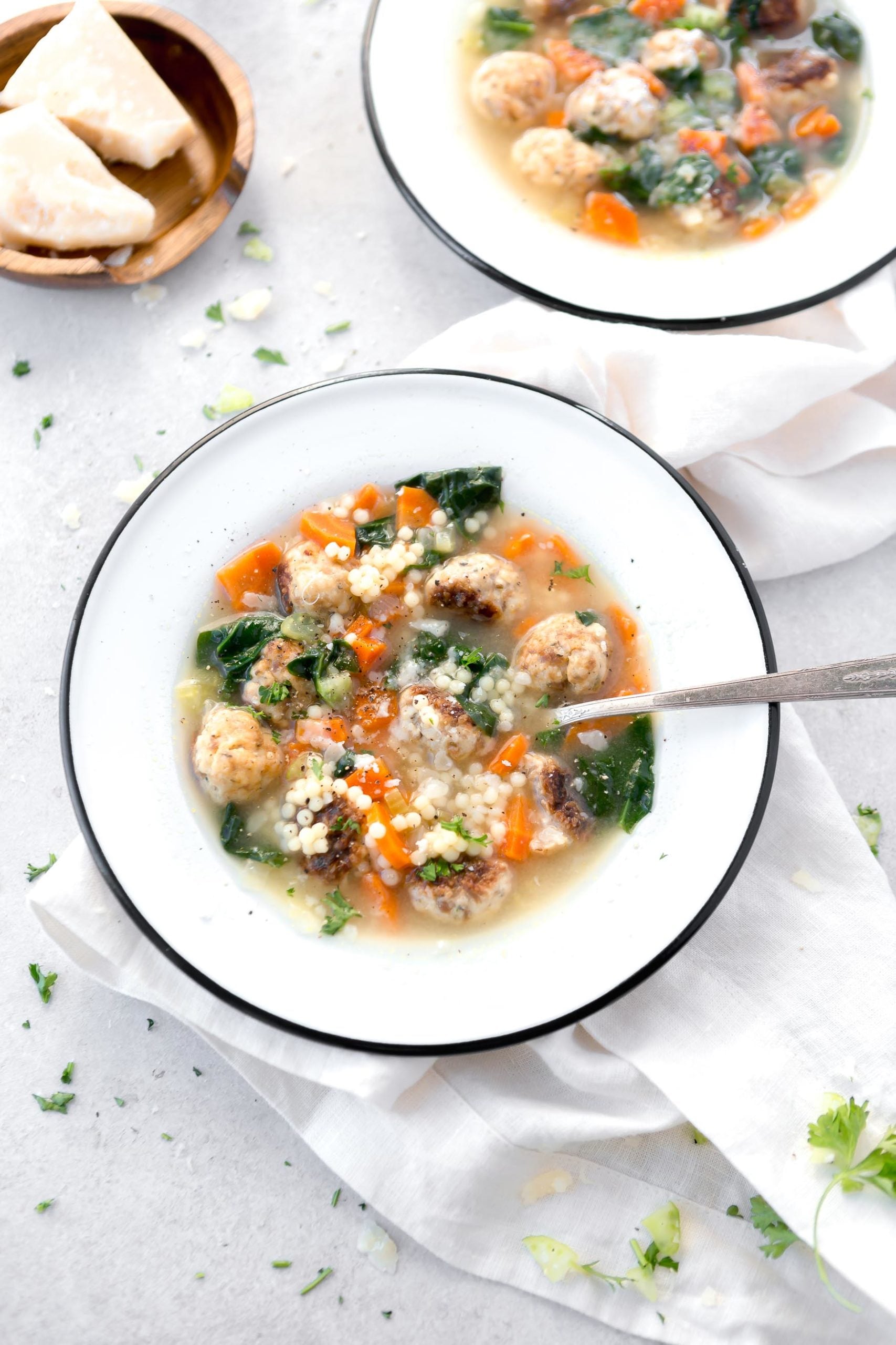 A bowl of Italian Wedding soup with a spoon in it. The soup is packed with colorful vegetables and meatballs. The bright green spinach brightens up the soup.