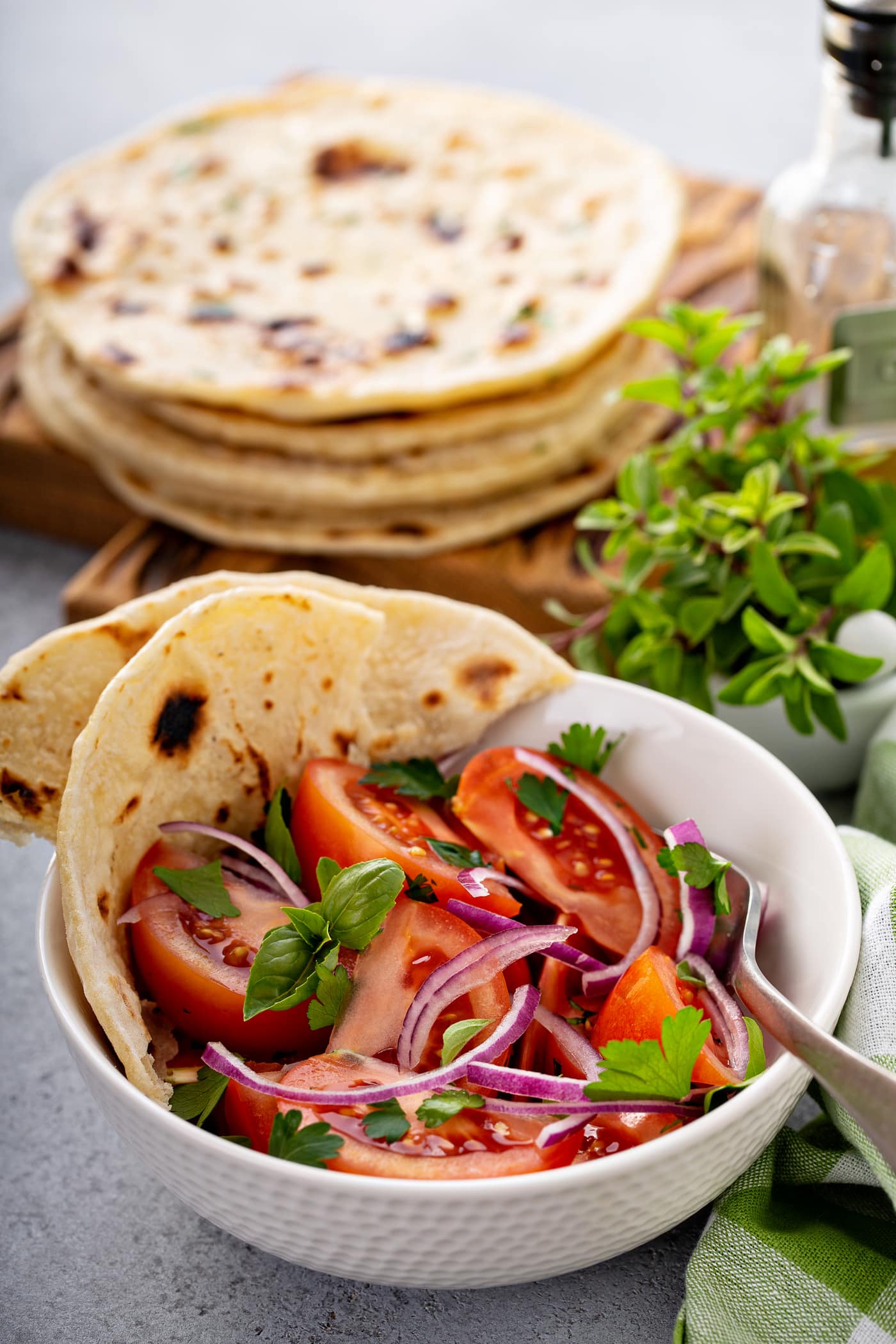 A photo of a bowl of fresh tomato and onion salad in a white bowl with torn pieces of unleavened bread sitting on the side of the bowl and a stack of cooked unleavened bread in the background.