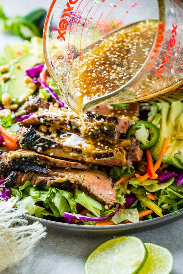 A grey plate full of mixed greens, veggies and thai peanut sauce marinated steak and a ginger dressing being poured over the top A grey plate full of mixed greens, veggies and thai peanut sauce marinated steak and a ginger dressing being poured over the top