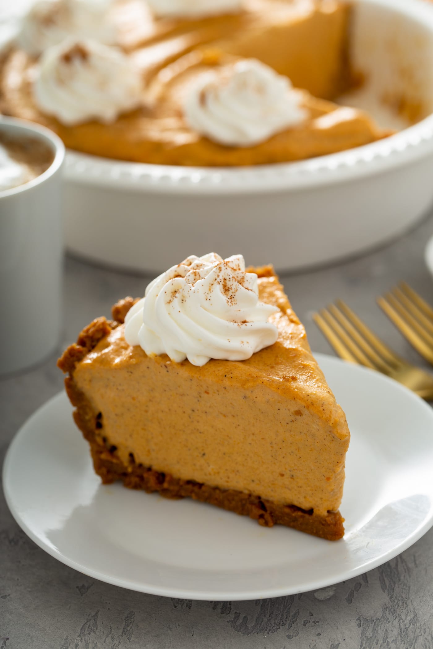 A piece of pumpkin ice cream pie on a white dessert plate. The pie has a dollop of whipped cream sprinkled with cinnamon on top. There are two forks beside the plate, and the rest of the pumpkin ice cream pie in a pie dish is in the background. 