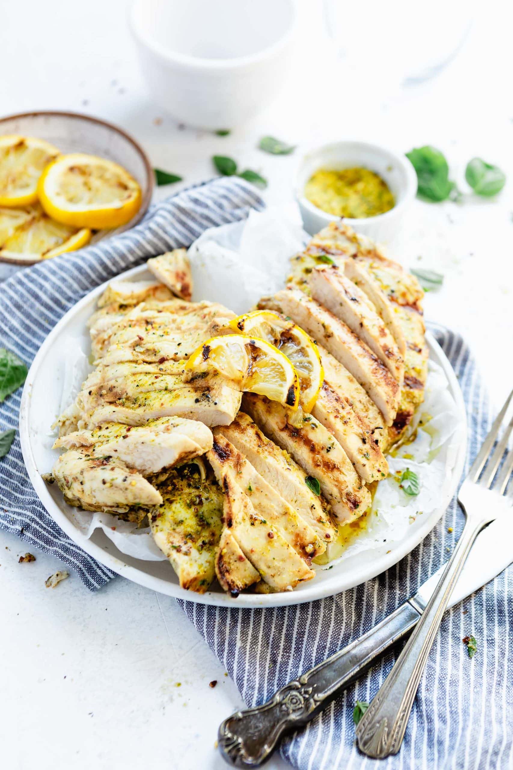 A plate of sliced, gilled chicken breasts with grilled lemon slices in the background. The plate is sitting on a blue and white striped cloth with a knife and fork next to the plate.