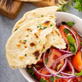 A photo of a bowl of fresh tomato and onion salad in a white bowl with torn pieces of unleavened bread sitting on the side of the bowl.