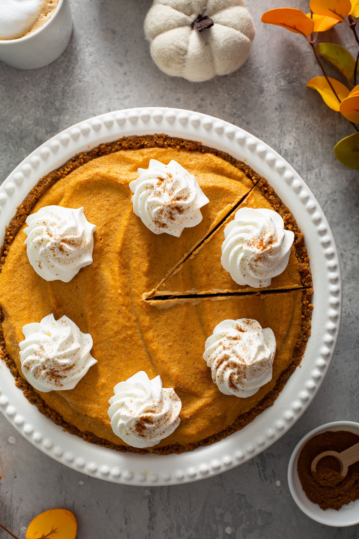 A whole pumpkin ice cream pie in a white pie dish. The pie has dollops of whipped cream sprinkled with cinnamon on top. A piece of pie has been cut but not removed from the dish. A small bowl of cinnamon, a tiny white pumpkin, and a mug of hot cocoa are in the background.