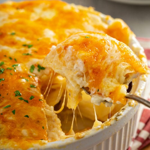 A casserole dish filled with cheesy potatoes. The cheese is melted to perfection and there are strings of cheesy hanging from the serving spoon.