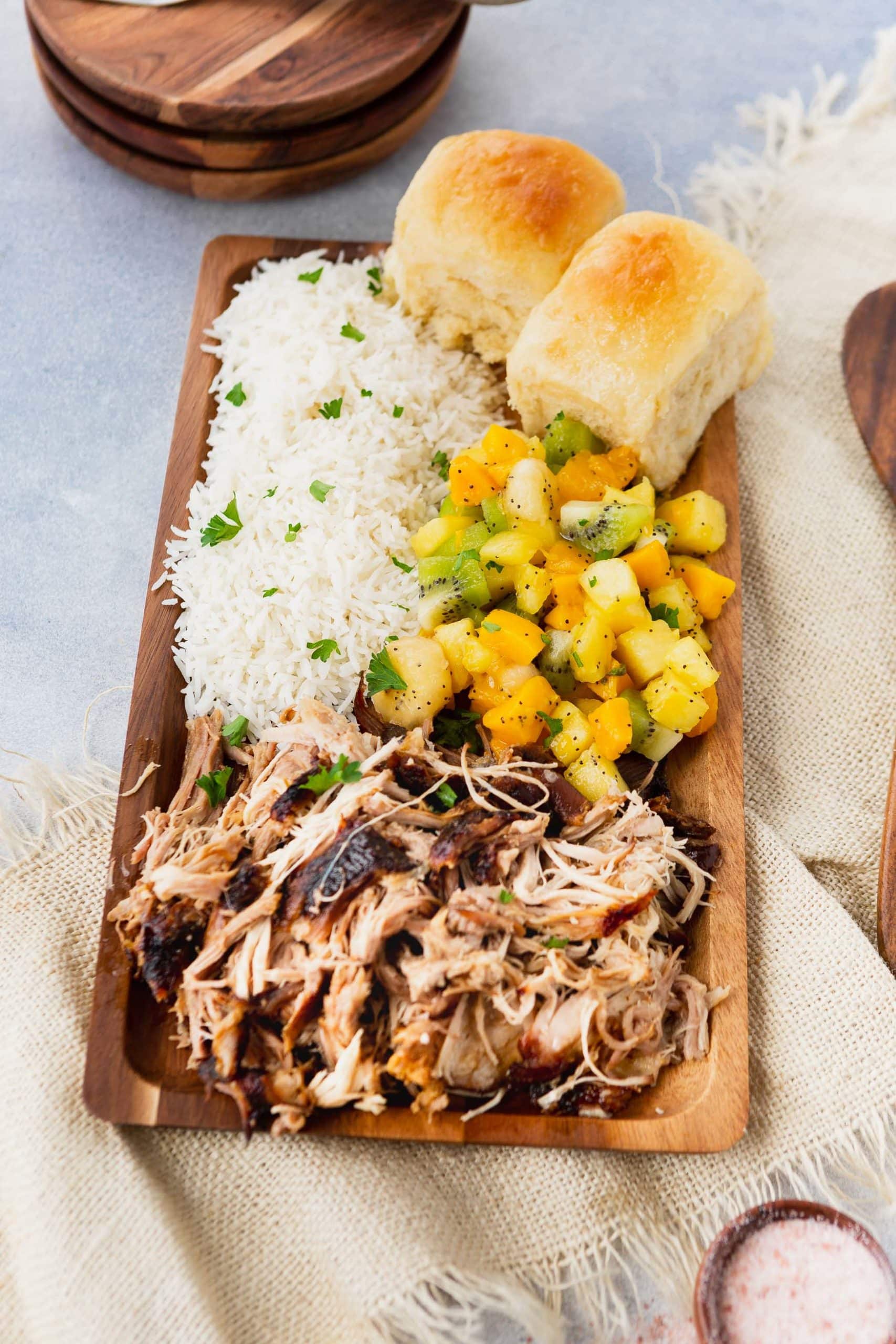 a photo of a rectangular wooden platter full of kalua pulled pork, herbed rice, tropical fruit salad and golden Hawaiian rolls.