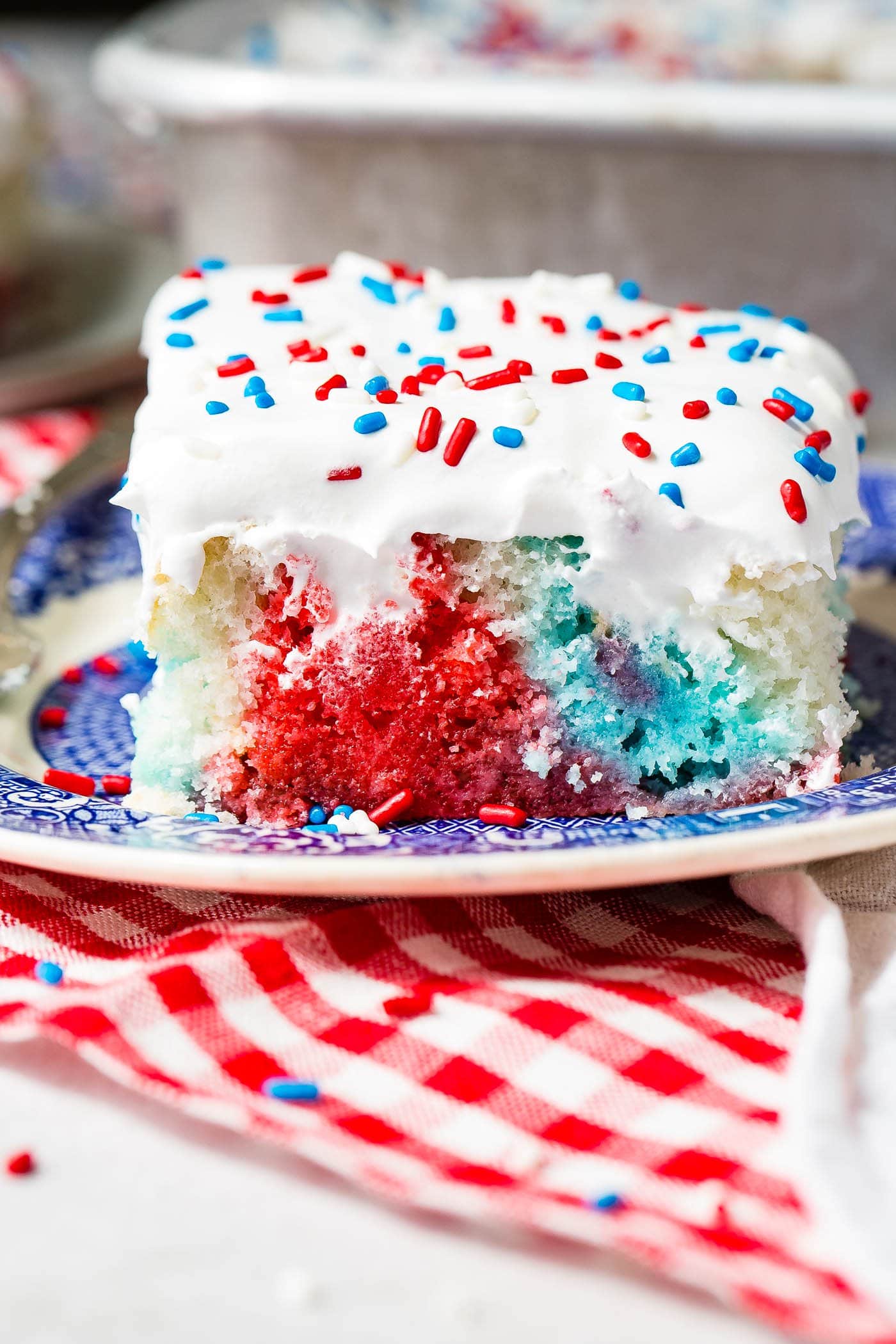 A piece of red, white and blue poke cake on a blue and white dessert plate. The cake is frosted with white frosting and sprinkled with red, white and blue sprinkles. You can see the red, white and blue portions of the cake where holes have been poked and jello poured in.