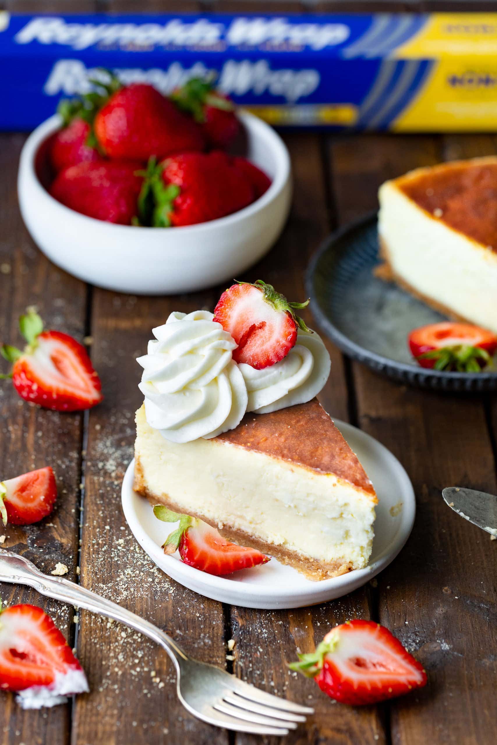 a photo of a slice of classic cheesecake topped with glazed whole strawberries and dollops of whipped cream on a small white dessert plate.