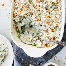 A casserole dish filled with green beans and feta. There is a serving spoon in the dish and you can see the creamy cheese sauce, green beans and sliced almonds on top. A dinner plate with a spoonful of beans is near the casserole dish.