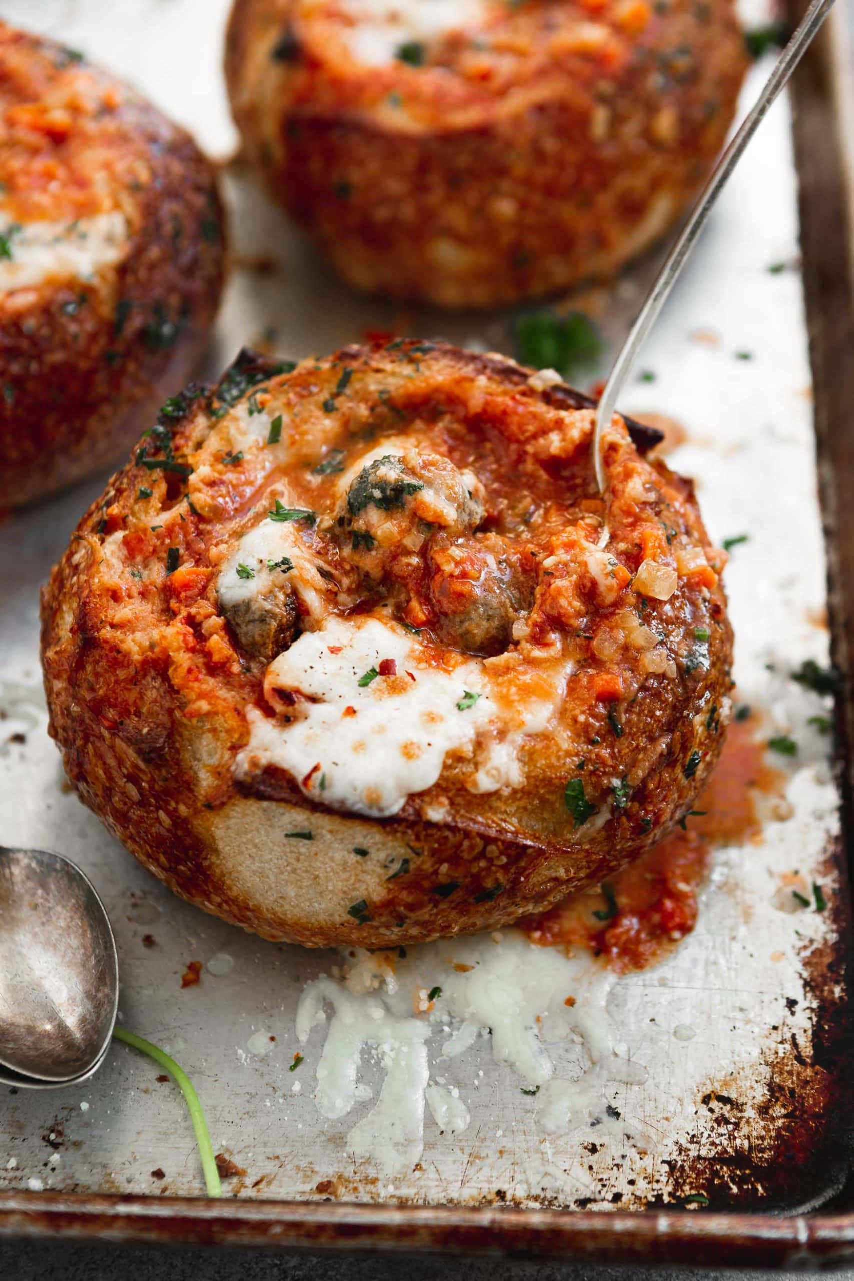 A bread bowl filled with parmesan meatball soup. The bread bowl and soup have been baked to a perfect toasty brown and the cheese on top is gooey melted perfection.
