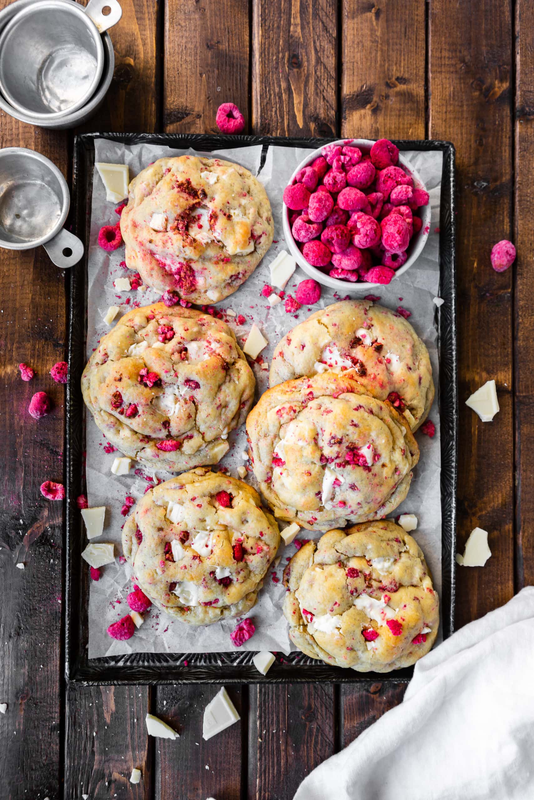 a photo of several large golden white chocolate raspberry cookies sitting on a white piece of parchment paper on top of a baking sheet with a cup of freeze dried raspberries sitting next to them and chunks of white chocolate scattered around them.