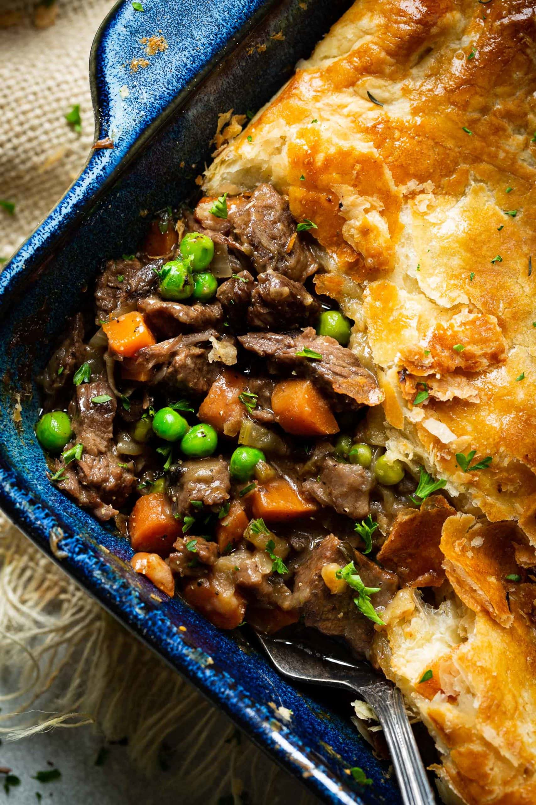 a photo of the corning of a baking dish full of beef pot pie with a serving removed so you can see the saucy beef filling with carrots and peas and onions and topped with golden puff pastry.