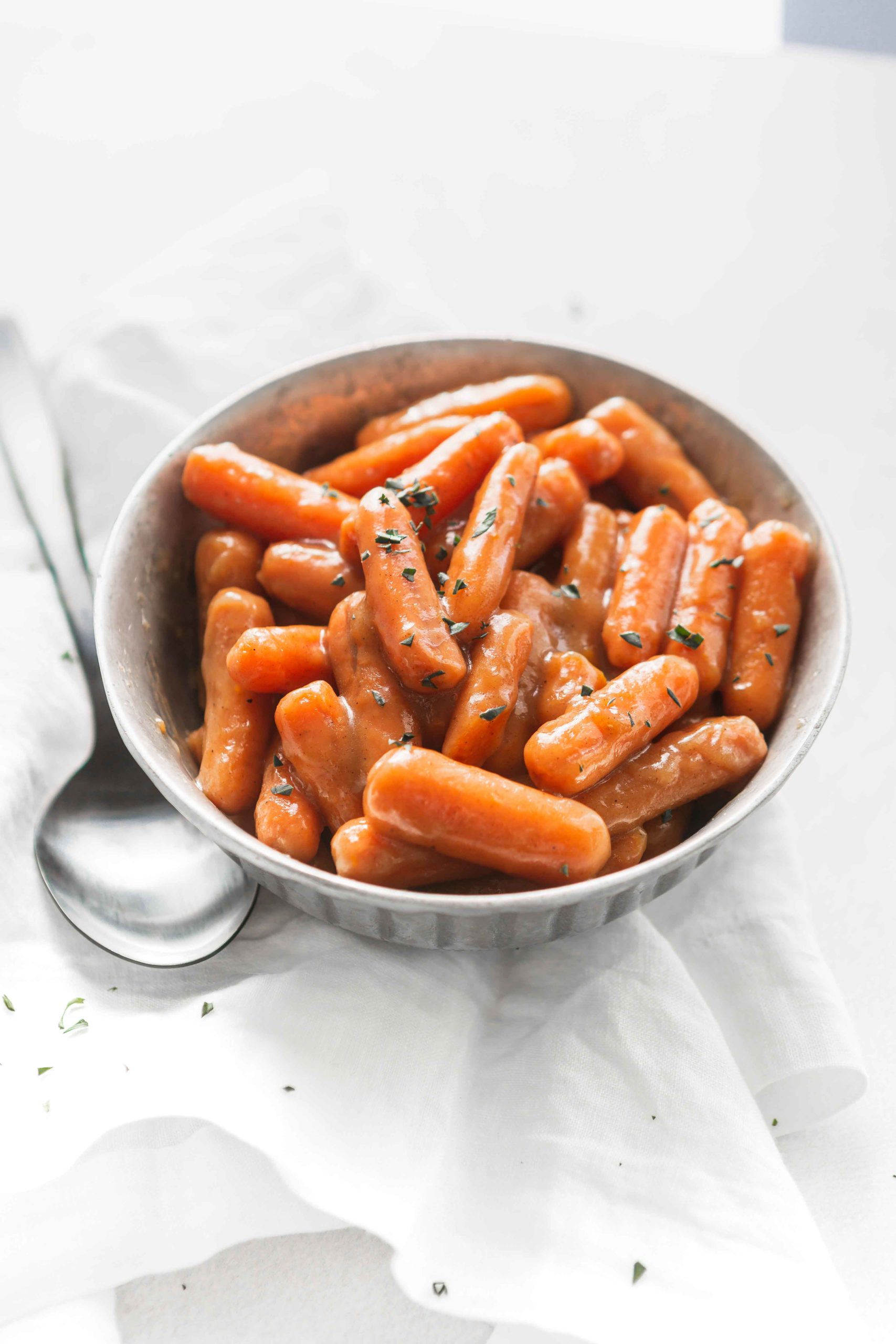 A bowl of orange glazed baby carrots with a little parsley sprinkled on top. A serving spoon is next to the bowl of carrots.