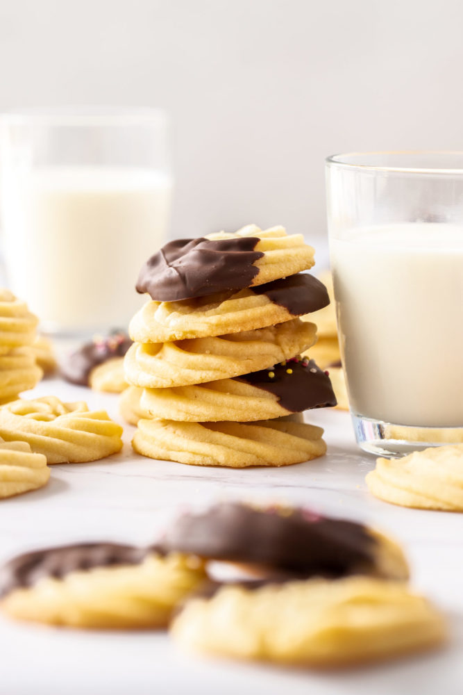A stack of butter cookies, some chocolate coated, sitting next to a glass of milk