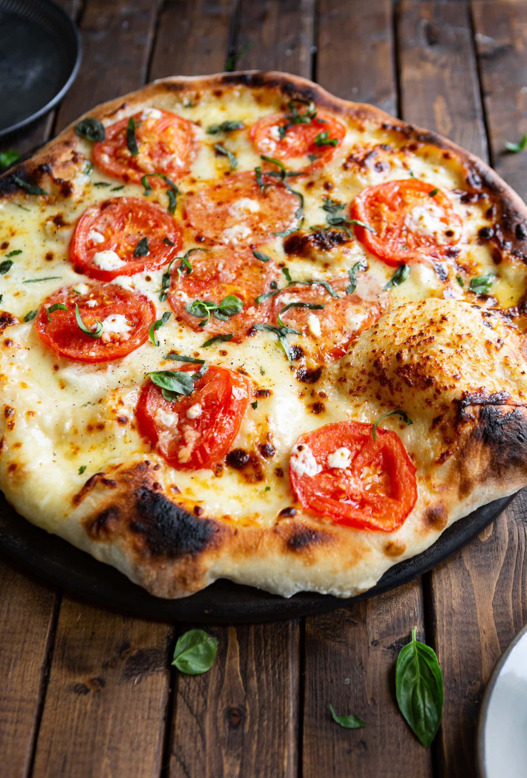 a photo of a whole thin crust margherita pizza topped with slices of tomato, chopped fresh basil and chunks of feta cheese.