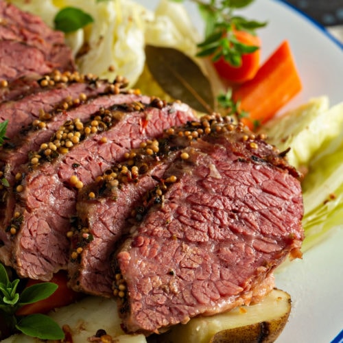 Slice corned beef resting on top of cabbage, potatoes and carrots with sprigs of herbs on the side.