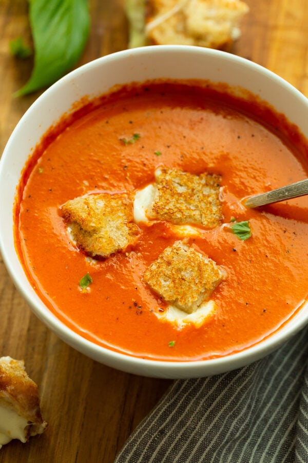 a photo of a bowl of tomato basil soup with three grilled cheese bites sitting on top and a spoon in the bowl