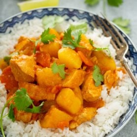 A bowl of white rice with chicken curry on top. there are chunks of chicken, carrots, and potatoes in a curry sauce. The curry is sprinkled with fresh parsley.