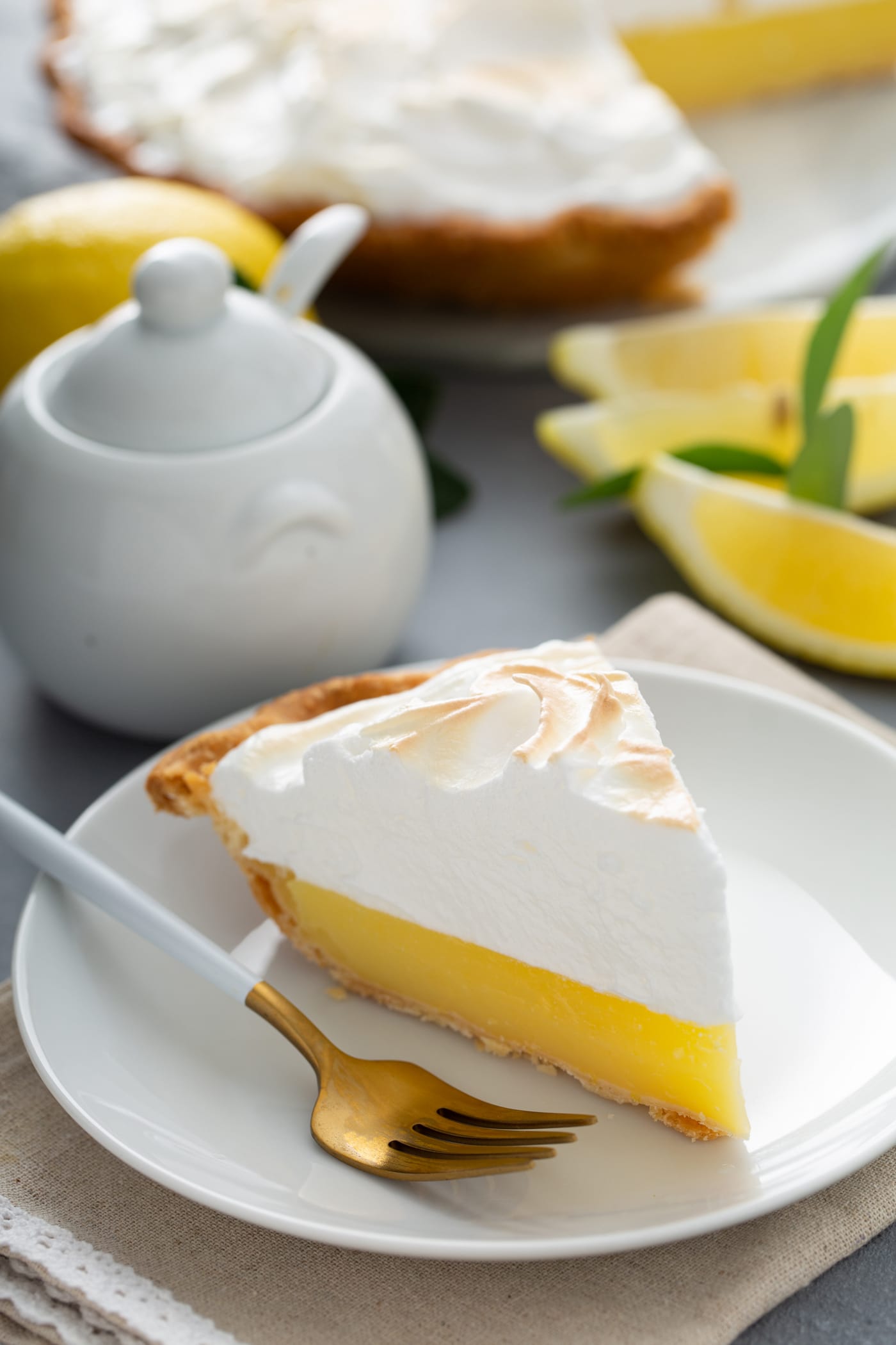 A picture of a slice of lemon meringue pie. The filling is bright yellow and the meringue on top is lightly toasted. There are lemon wedges and a sugar bowl in the background and a fork is on the plate next to the slice of pie.