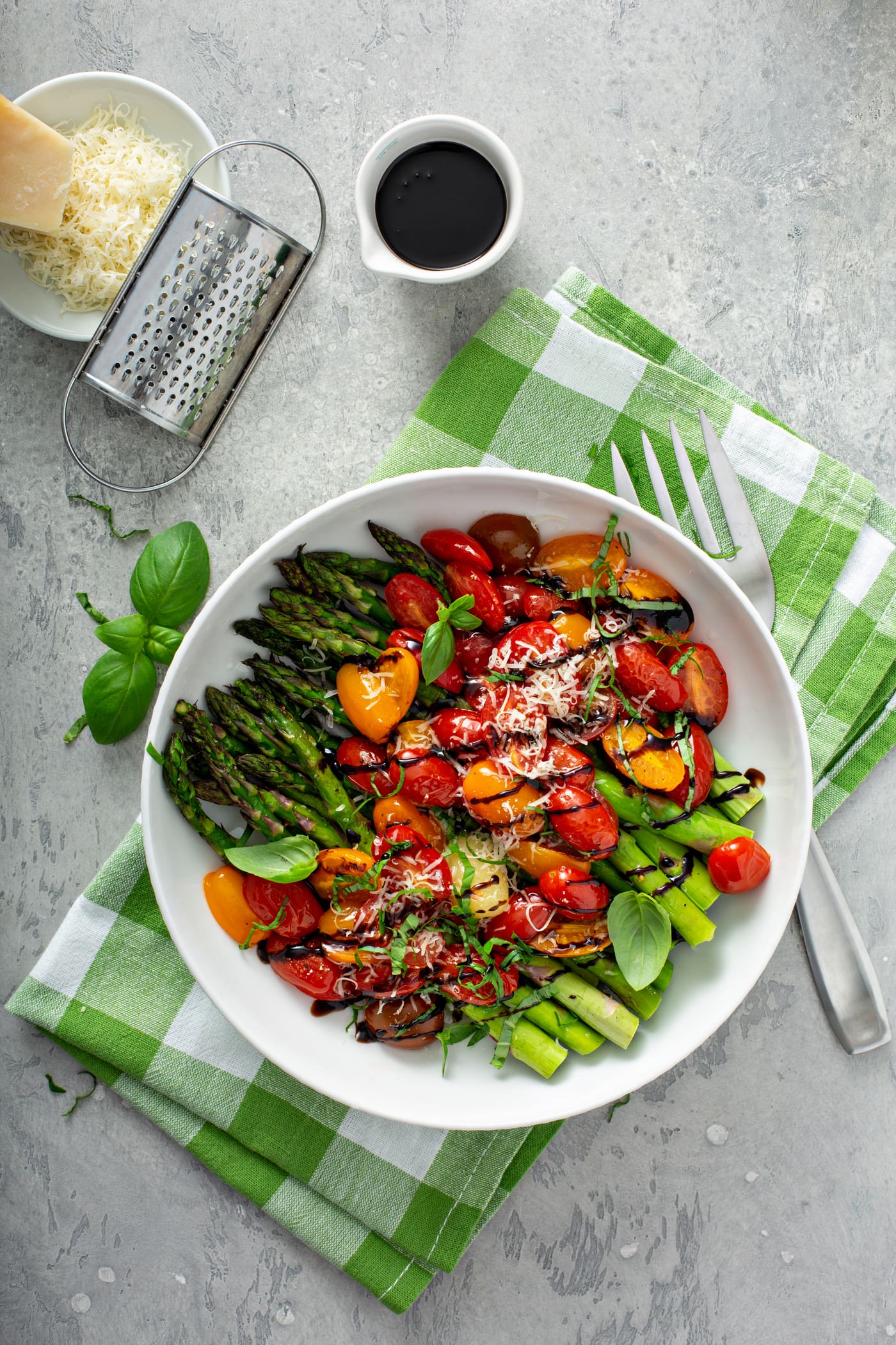 A serving dish of balsamic glazed tomatoes and asparagus. The bright green asparagus and sliced red and yellow heirloom tomatoes are drizzled with balsamic glaze and sprinkled with grated Parmesan cheese. The serving dish is sitting on a green and white checked cloth. There is a fork beside the dish and a cup of balsamic glaze and a dish of grated cheese and a cheese grater in the background.