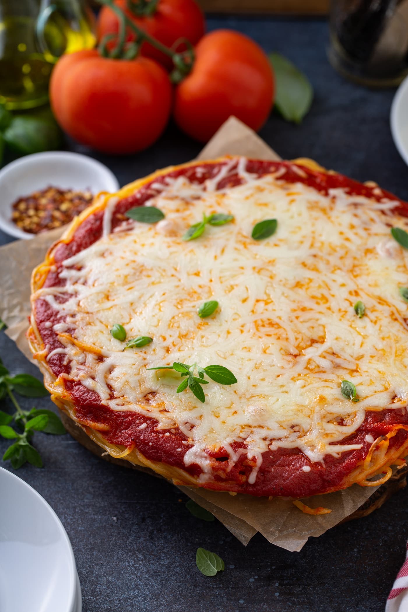 A baked spaghetti pie in a pie dish. There are layers of spaghetti, sauce, and melted cheese. Fresh basil is sprinkled on top and three tomatoes, olive oil, a container of dried red pepper flakes, and dinner plates are next to the spaghetti pie.