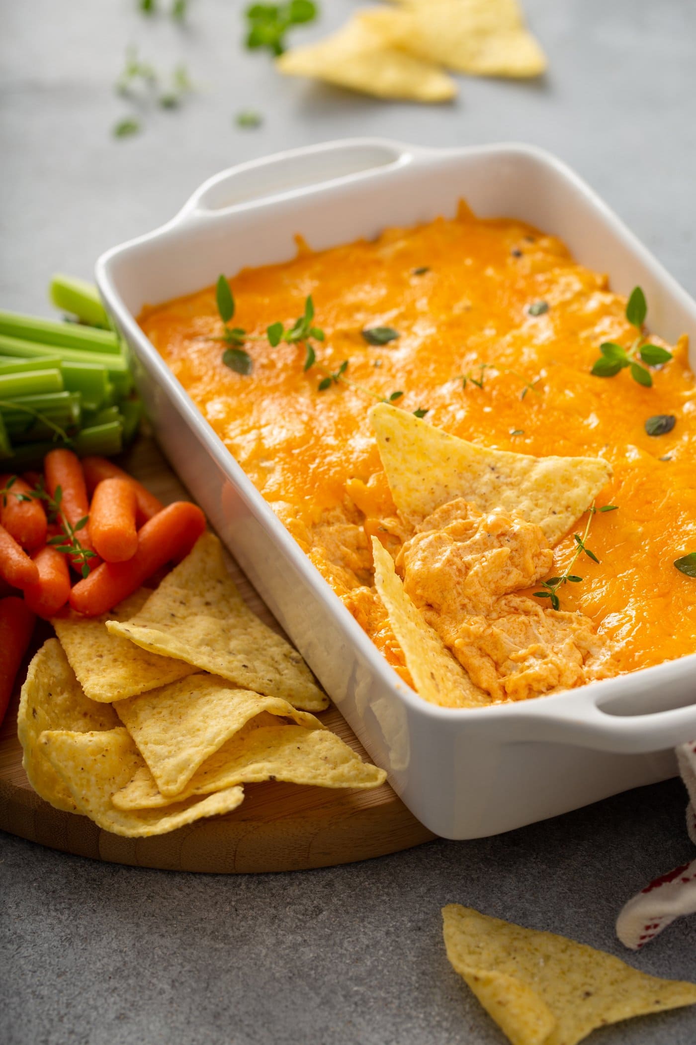 A casserole dish with buffalo chicken dip. A scoop has be taken out of the dip and there are a couple of chips laying in the dip. The dip is covered in melted cheese and sprinkled with parsley leaves. There are chips, carrots and celery laying next to the dip.