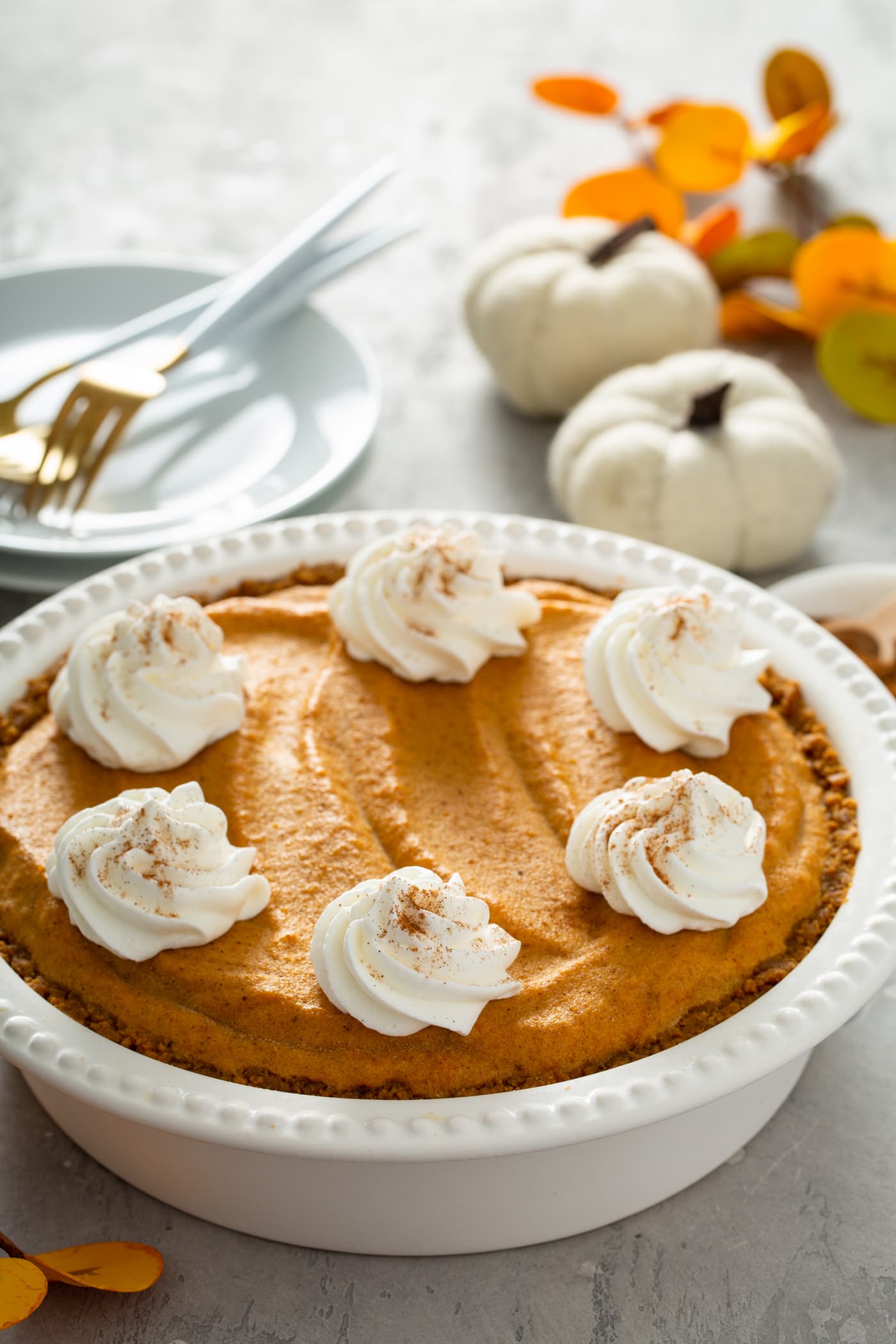 A whole pumpkin ice cream pie in a white pie dish. The pie has dollops of whipped cream sprinkled with cinnamon on top. There is a stack of white dessert plates with three forks on them, two tiny white pumpkins, and fall colored leaves behind the pie.