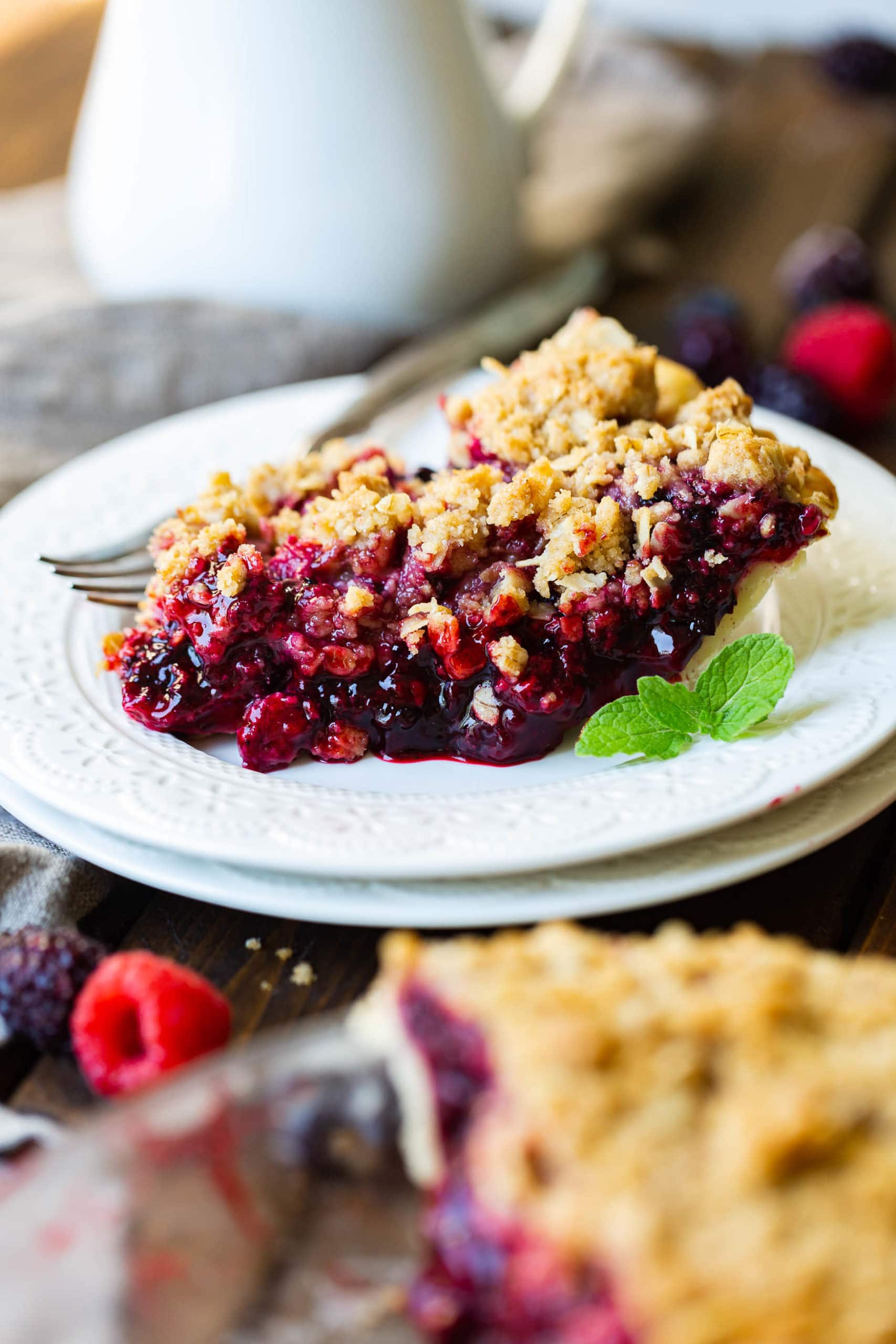a photo of a single slice of mixed berry pie with streusel topping sitting on a white desset plate with a sprig of mint sitting next to it.