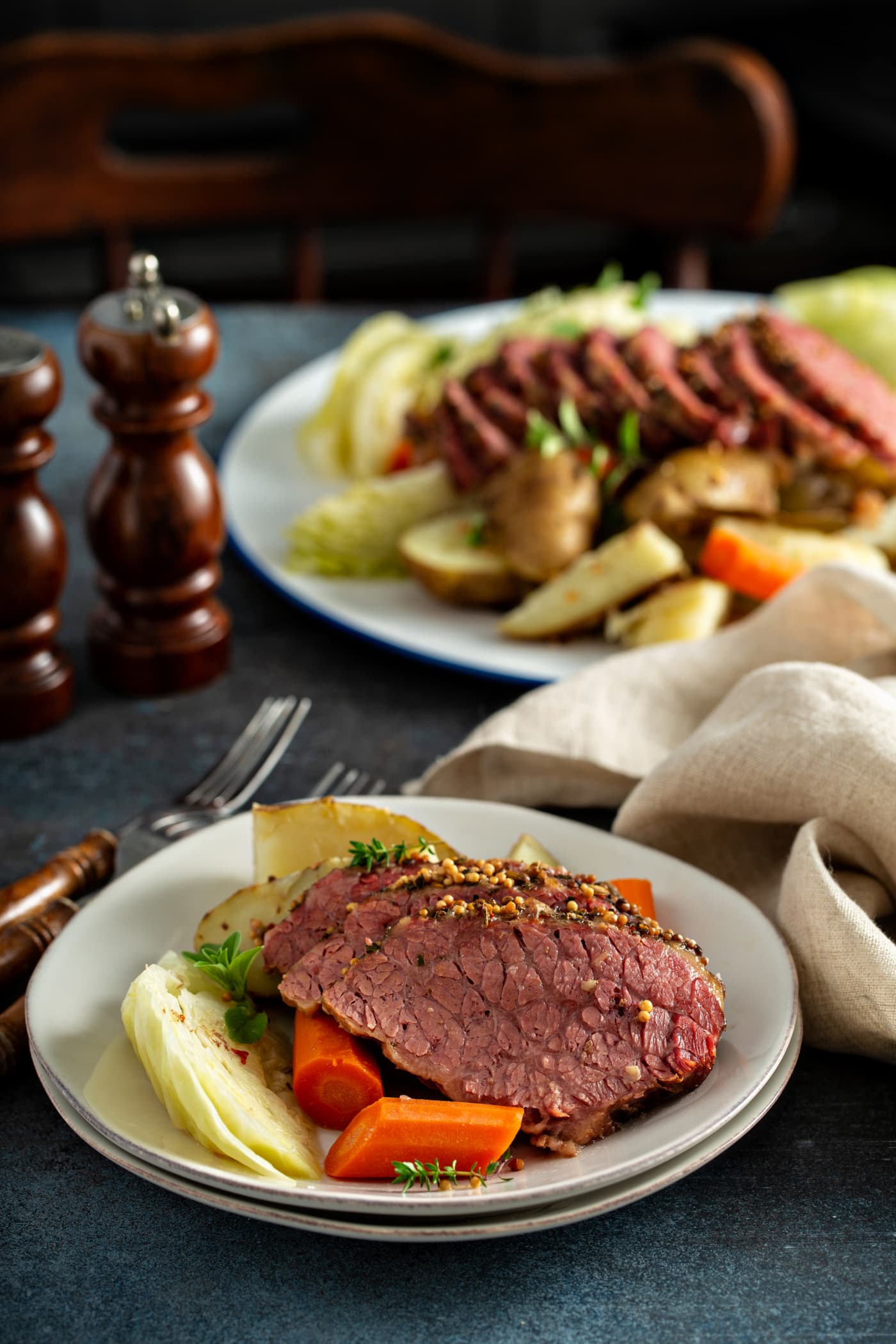 A dinner table with a platter of sliced corned beef and cabbage surrounded by potatoes, carrots, and cabbage. There is a dinner plate with three slices of corned beef and carrots, potatoes and cabbage. There are two forks on the table next to the plate and a wooden salt and pepper shaker in the background. There is also a tan linen napkin bunched up by the plate.