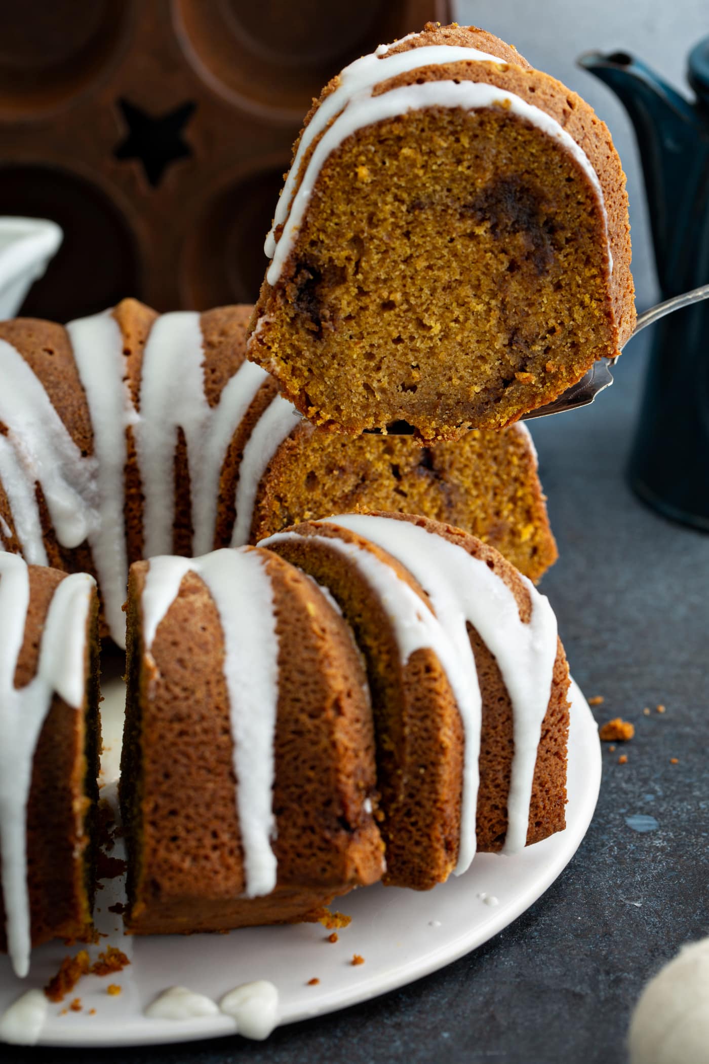A sour cream cinnamon swirl pumpkin bundt cake that has a drizzle of glaze on top. A slice of cake is being removed with a cake server. You can see a rich cinnamon swirl in the middle of the cake.