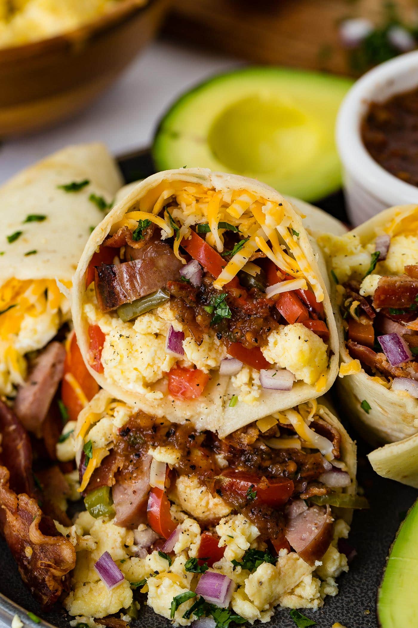 Apple chicken sausage burritos. The are stuffed with sausage, bacon, scrambled eggs, cheese, and peppers. There is a bowl of salsa and an avocado that is cut in half in the background.