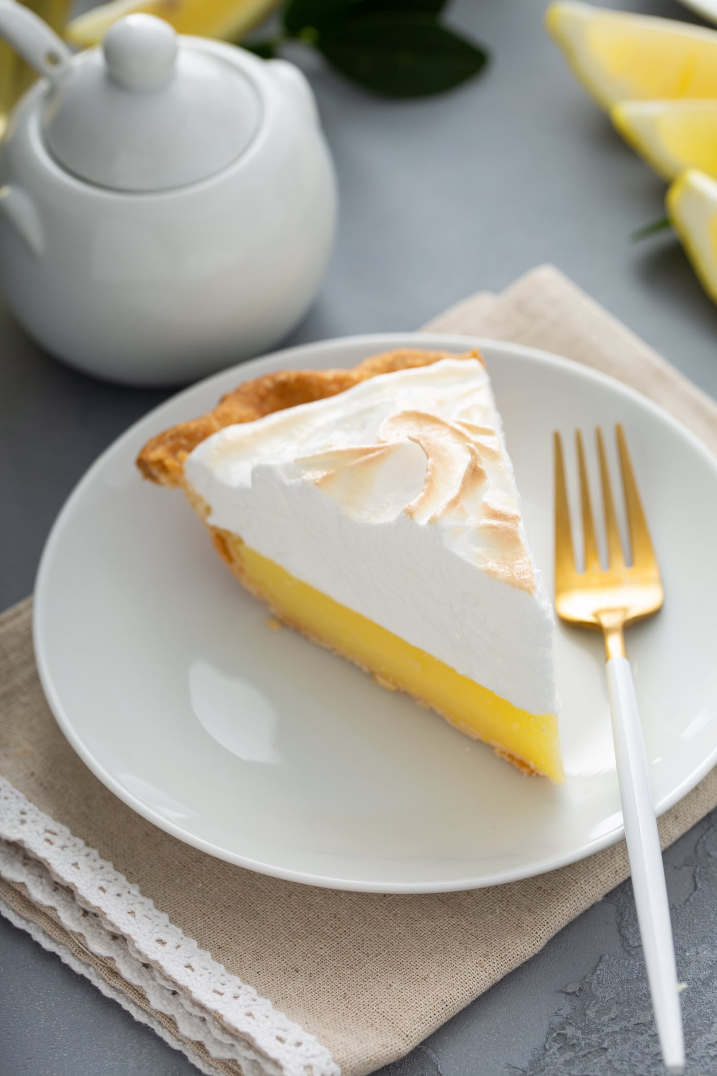 A slice of lemon meringue pie on a white dessert plate with a fork next to it. A sugar bowl is in the background.