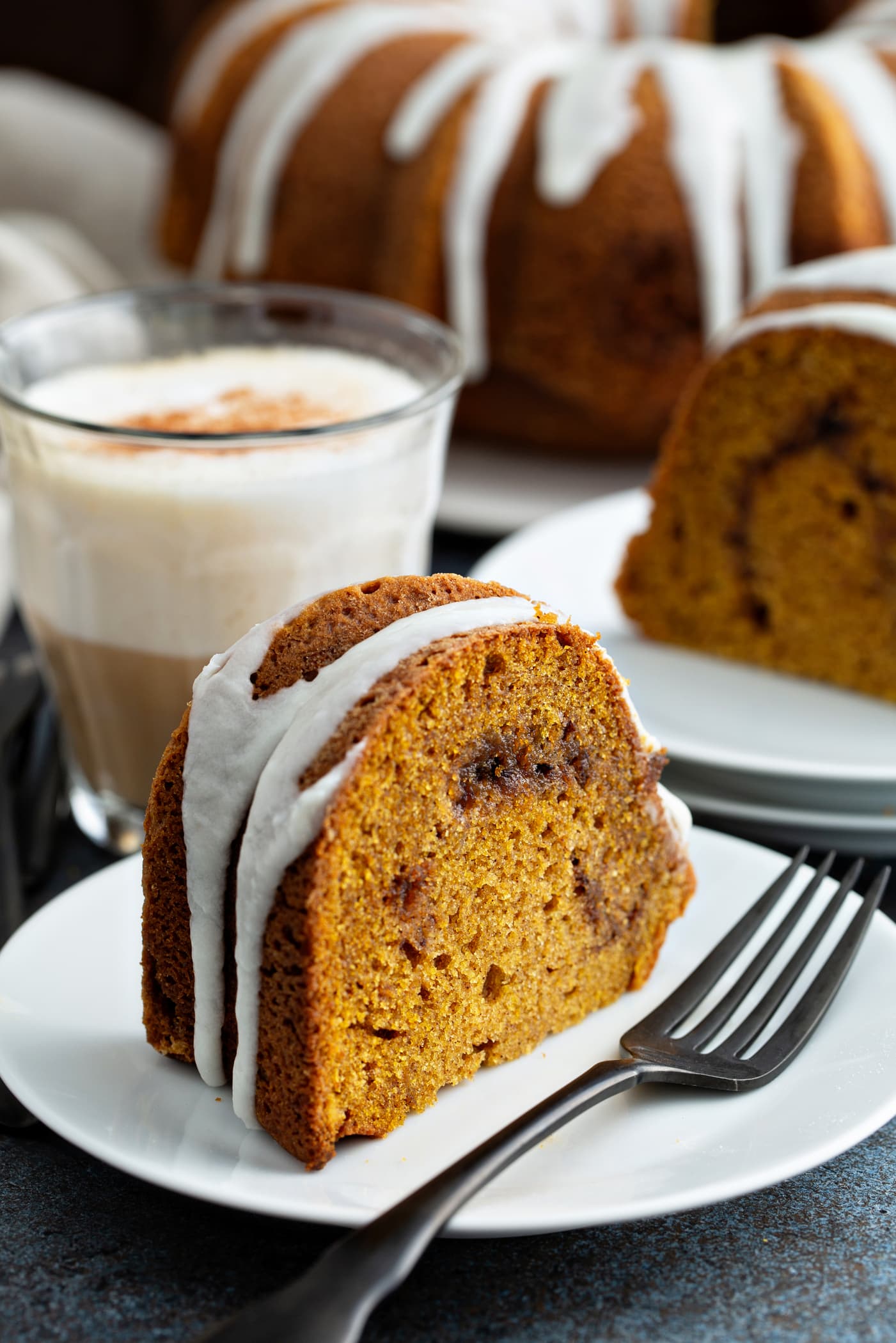 A slice of sour cream cinnamon swirl pumpkin bundt cake on a dessert plate with a fork on the plate next to the cake. The cake has a drizzle of white glaze on top. another slice of cake on a dessert plate, and the rest of the bundt cake on a cake platter are in the background.