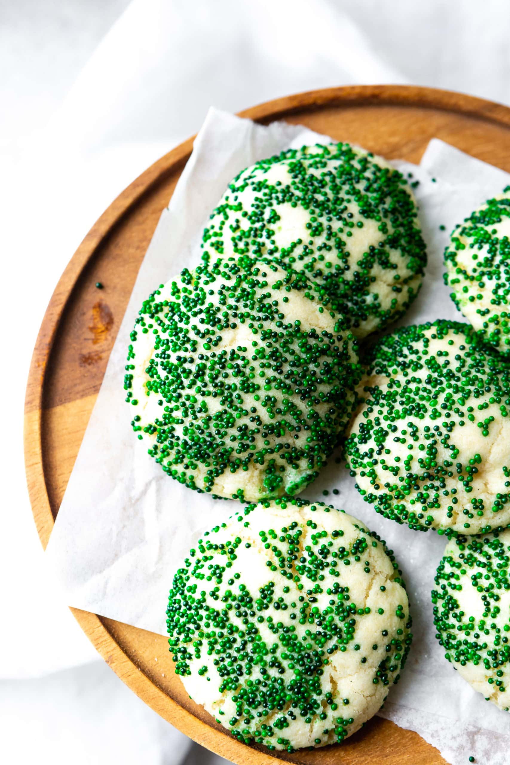 a photo of several thick and soft sugar cookies sitting on a piece of white parchment paper on top of a wooden serving tray. the cookies are all coated in dark green sprinkles.