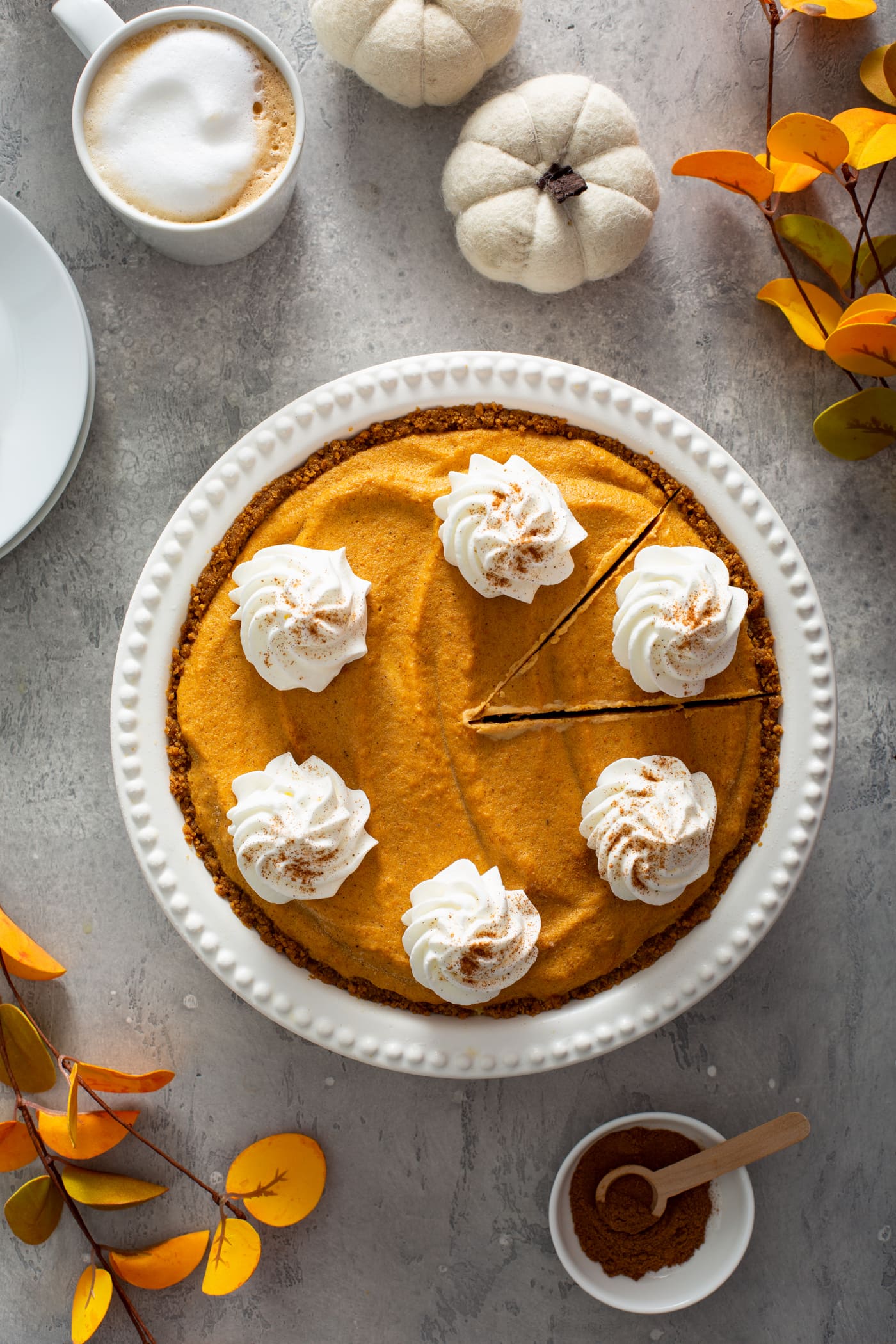 A whole pumpkin ice cream pie in a white pie dish. The pie is topped with dollops of whipped cream sprinkled with cinnamon. A slice has been cut but not removed from the pie. There is a dessert plate, a container of whipped cream, Two tiny white pumpkins, fall colored leaves and a small bowl of cinnamon in the background.