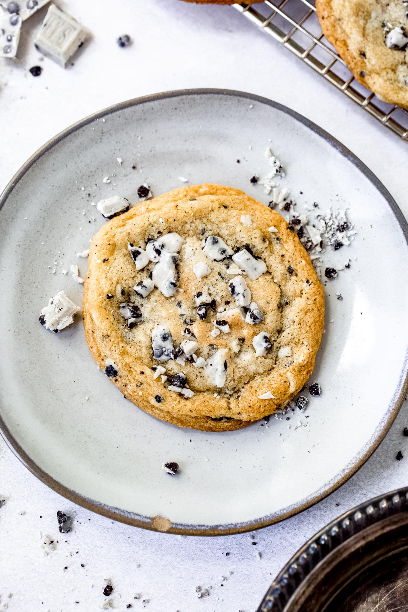 A dessert plate holding a large cookies and cream cookie. The cookie is loaded with bits of cookies and cream candy bar and there are bits of candy bar on the plate.
