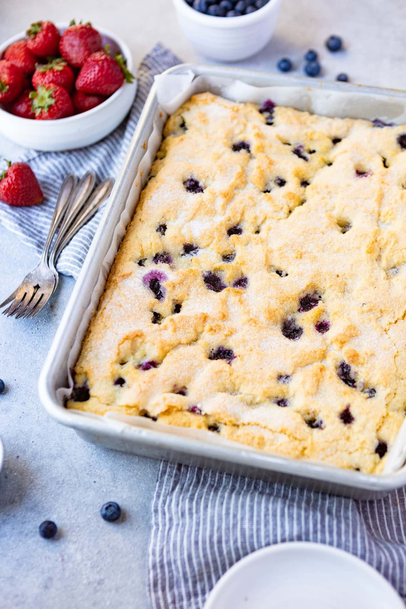 a photo of a golden brown blueberry breakfast cake in a 9x13 silver cake pan with two forks sitting along side the pan.
