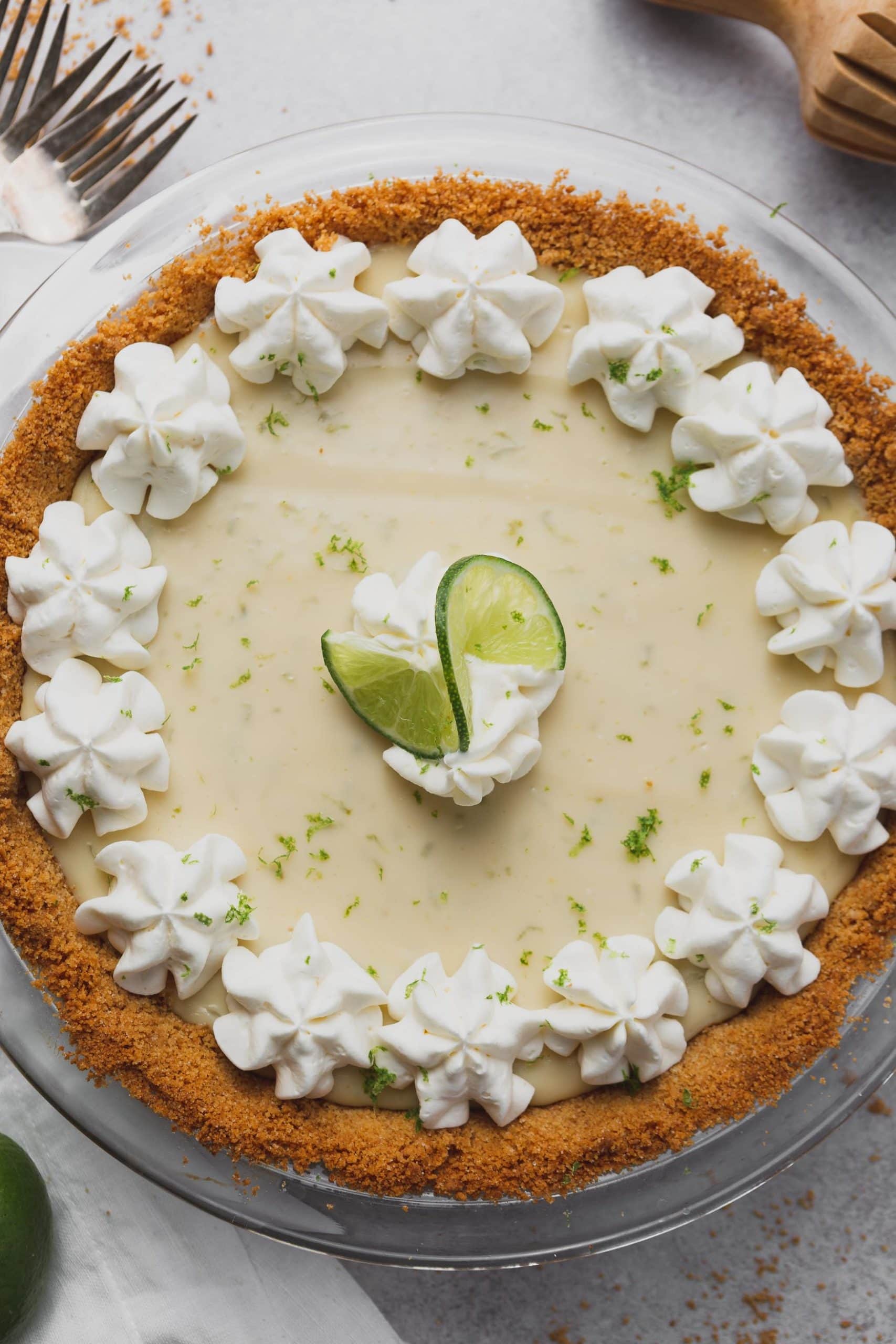 a top view of a key lime pie in a graham cracker crust. The pie has bits of lime zest, dollops of whipped cream and a curl of lime on top.
