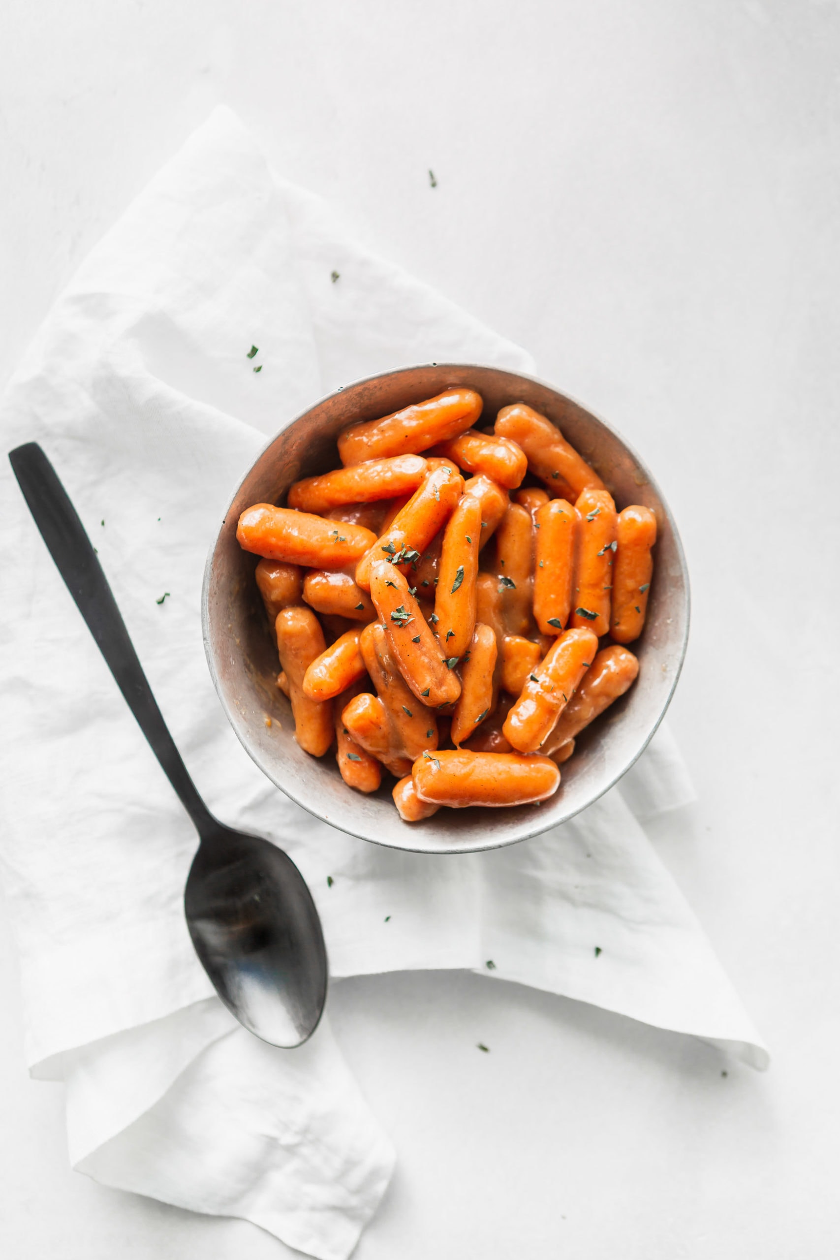 A serving bowl of orange glazed baby carrots. There are a few herbs sprinkled on top and a serving spoon is next to the bowl.
