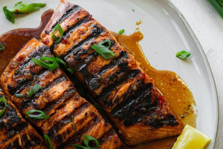 two grilled salmon filets sitting in a marinade sauce on a white plate and topped with chopped green onions