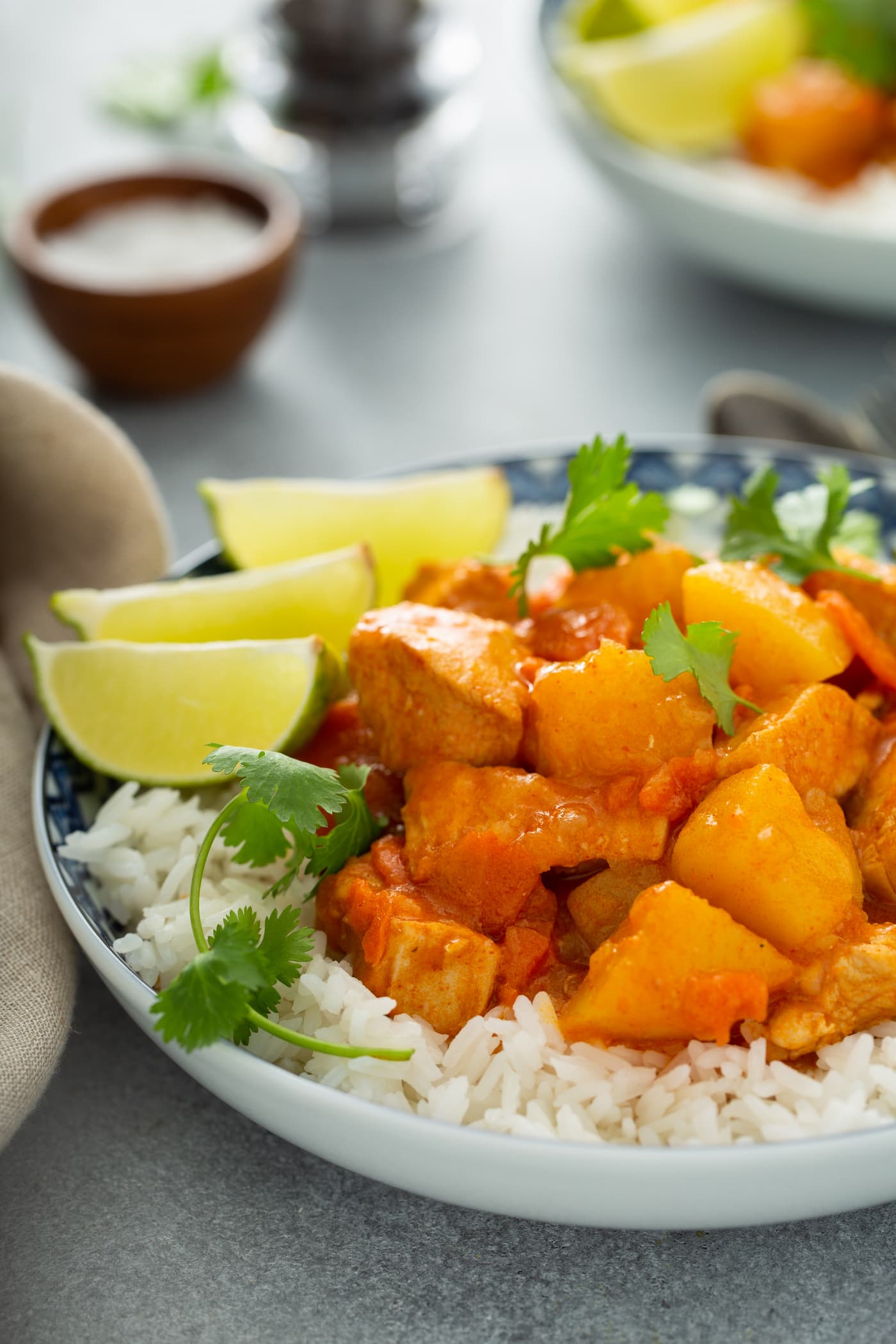 A bowl of white rice topped with chicken curry. There are chunks of chicken, potatoes and carrots in a curry sauce. It is sprinkled with fresh parsley and there are three lime wedges on the edge of the bowl. A container of salt and pepper are in the background.