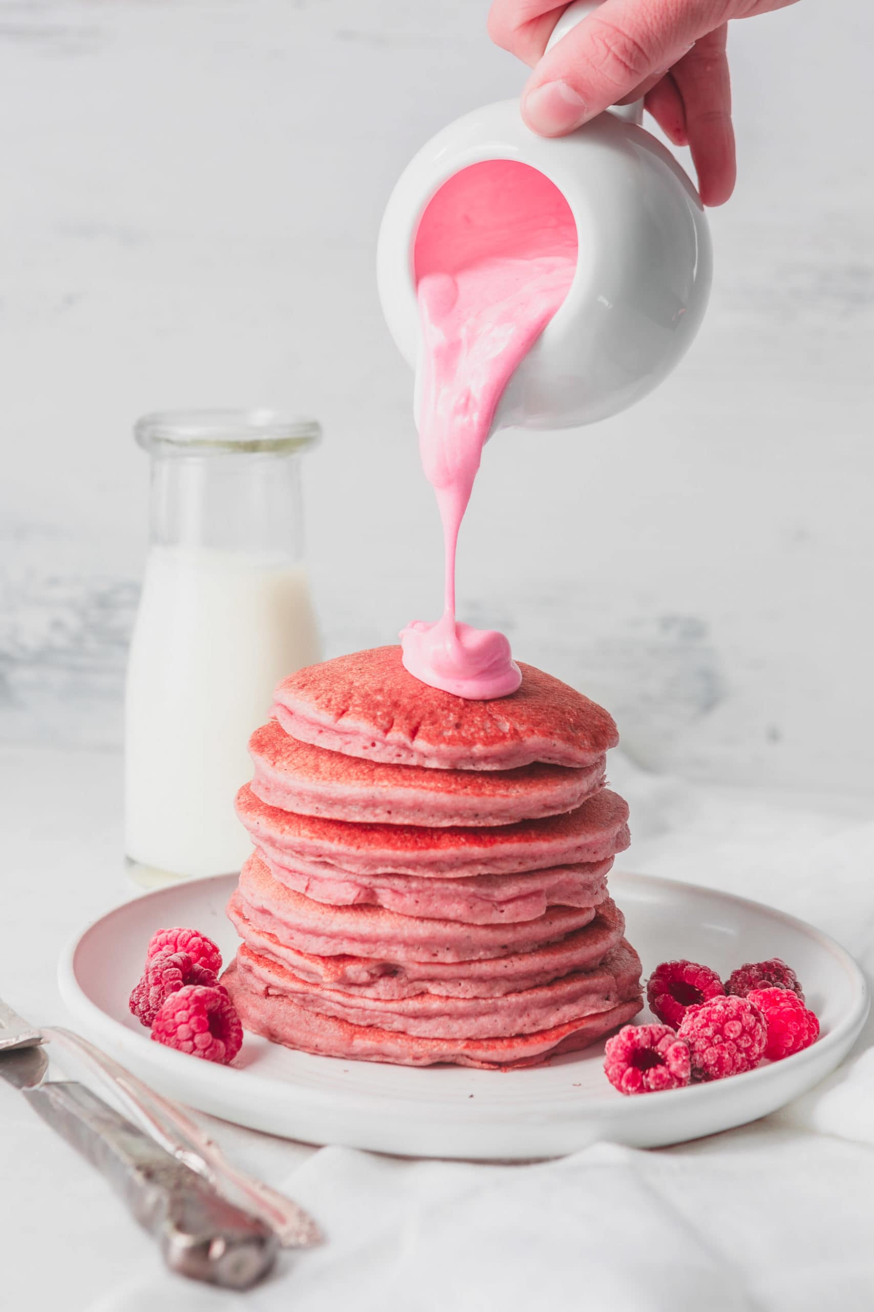 A stack of pink pancakes with a thick pink chocolate sauce being poured over the top. There are fresh raspberries on the plate with the pancakes and a container of milk in the background. with 