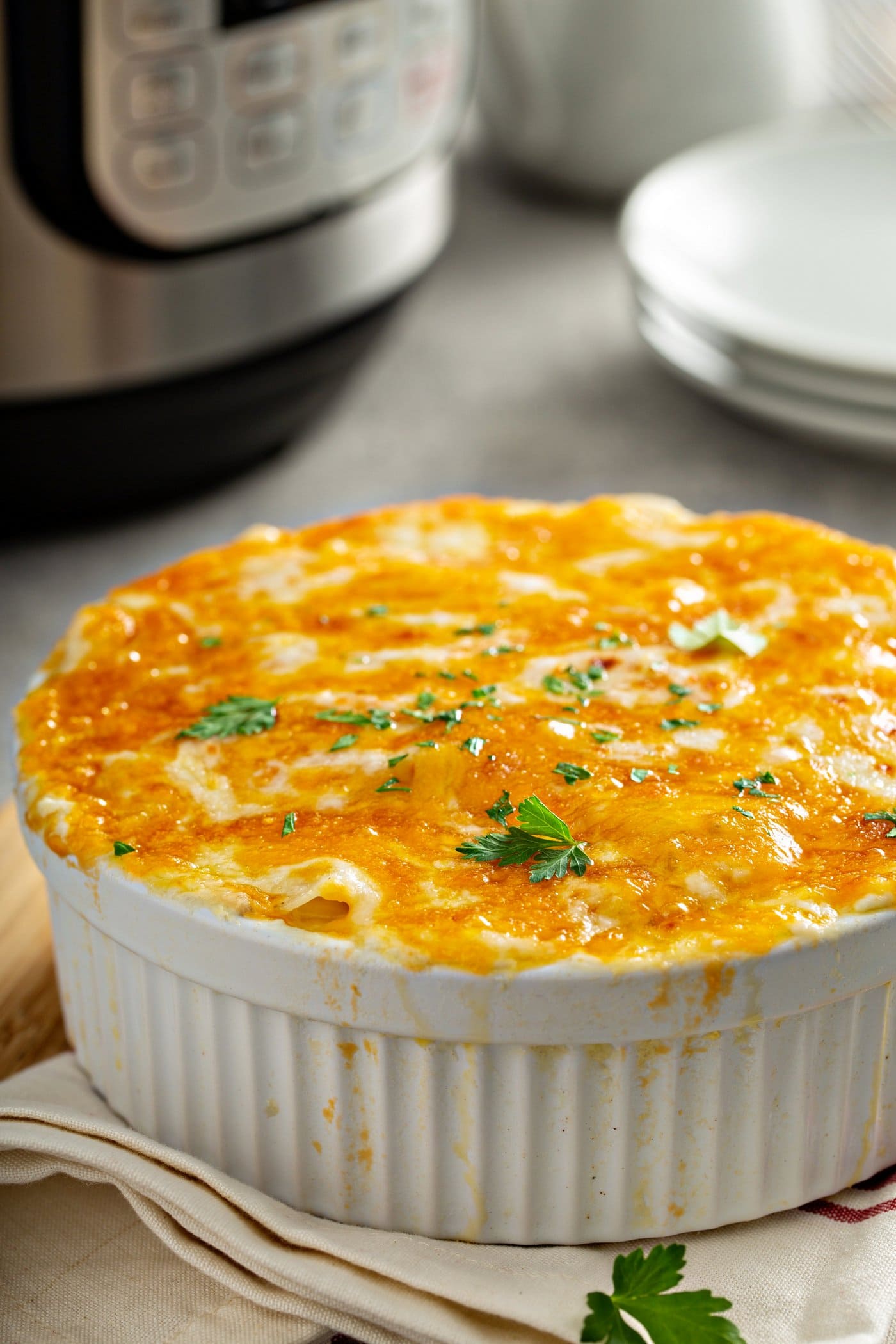 A casserole dish with cheesy potatoes topped with melted cheese and parsley leaves. There is a stack of dinner plates in the background and an instant pot.