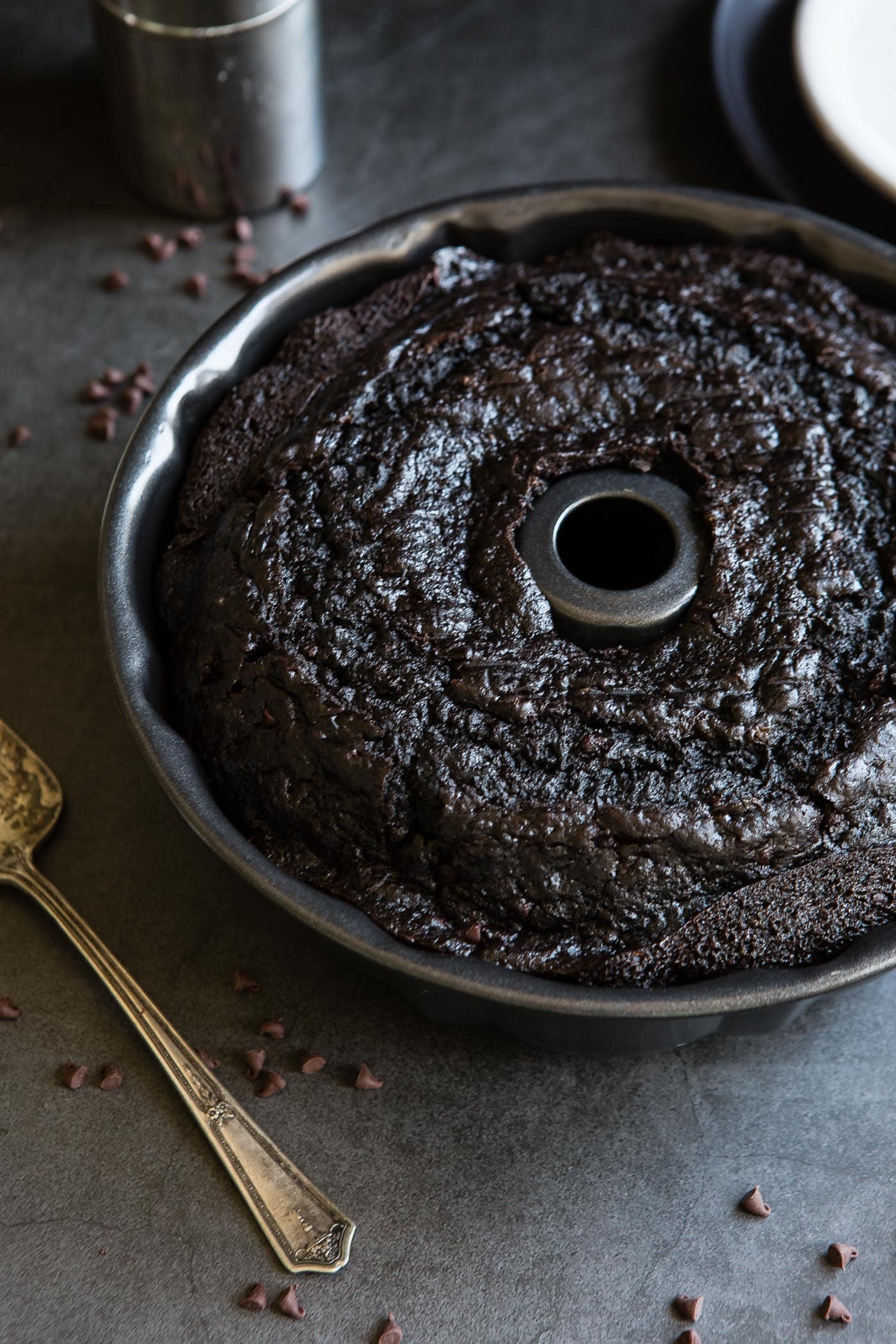A baked zucchini chocolate bundt cake in the bundt pan. Bits of chocolate and a cake server are next to the cake. 
