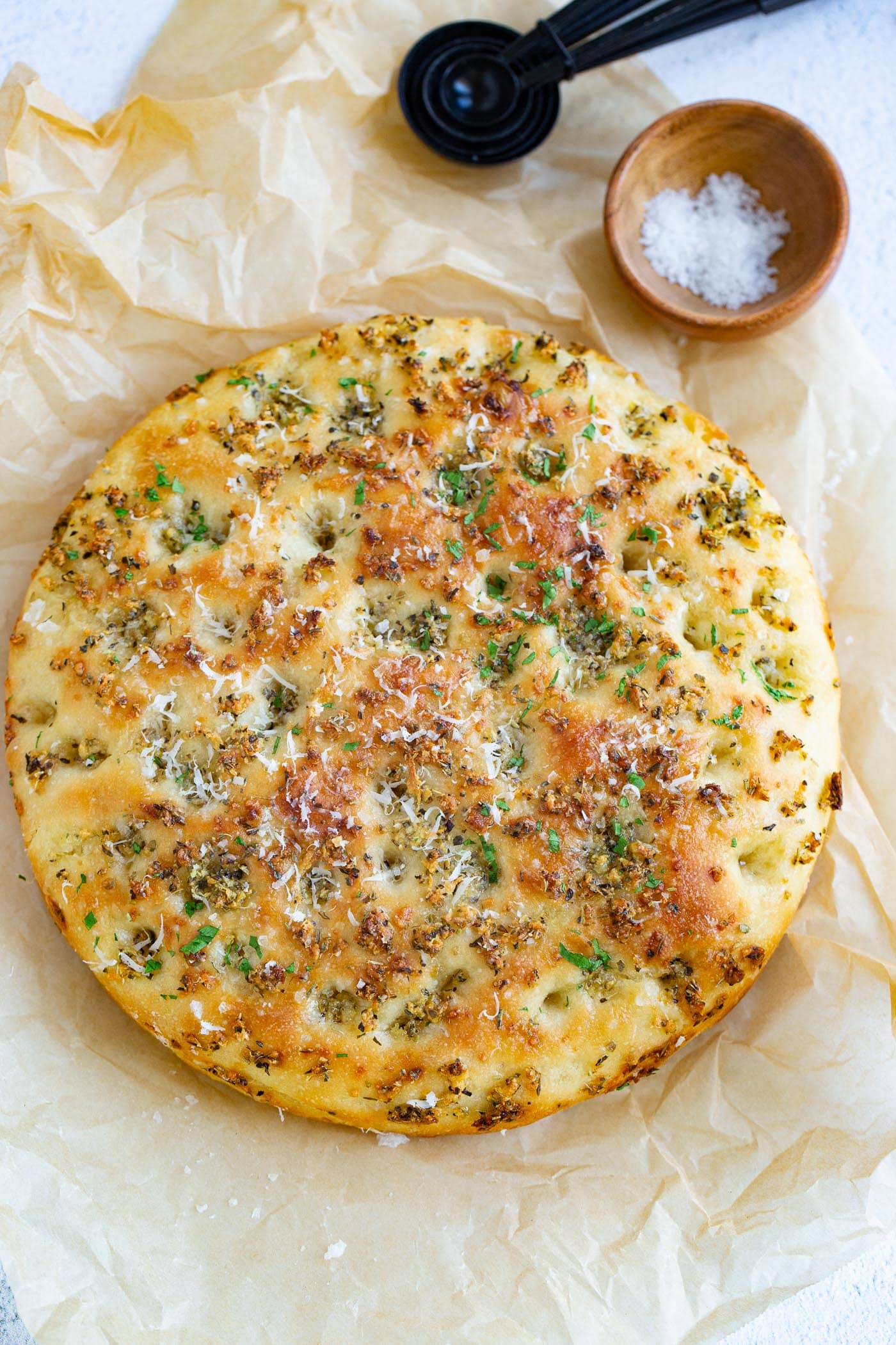 Baked garlic parmesan focaccia bread sitting on parchment paper. It is baked to a golden brown with bits of green herbs on it. There is a small container of salt in the background.