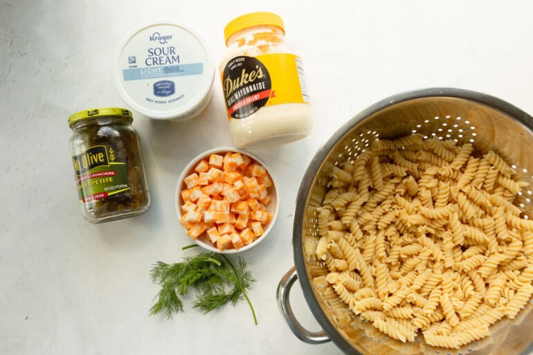 A colander filled with rotini pasta, dill, pickles, cubes of cheese, dukes mayo, and sour cream.