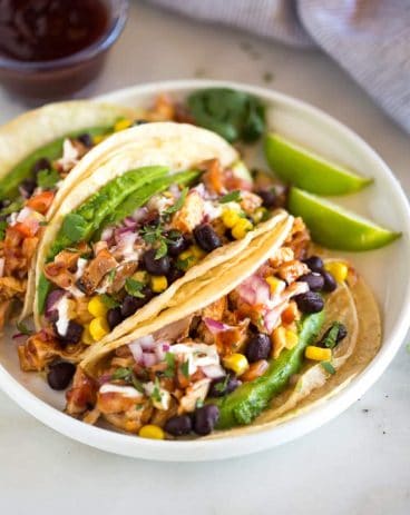 Grilled BBQ Chicken Tacos topped with corn, black beans, avocado, onion and tomatoes, served on a white plate with a small container of extra bbq sauce in the background Grilled BBQ Chicken Tacos topped with corn, black beans, avocado, onion and tomatoes, served on a white plate with a small container of extra bbq sauce in the background